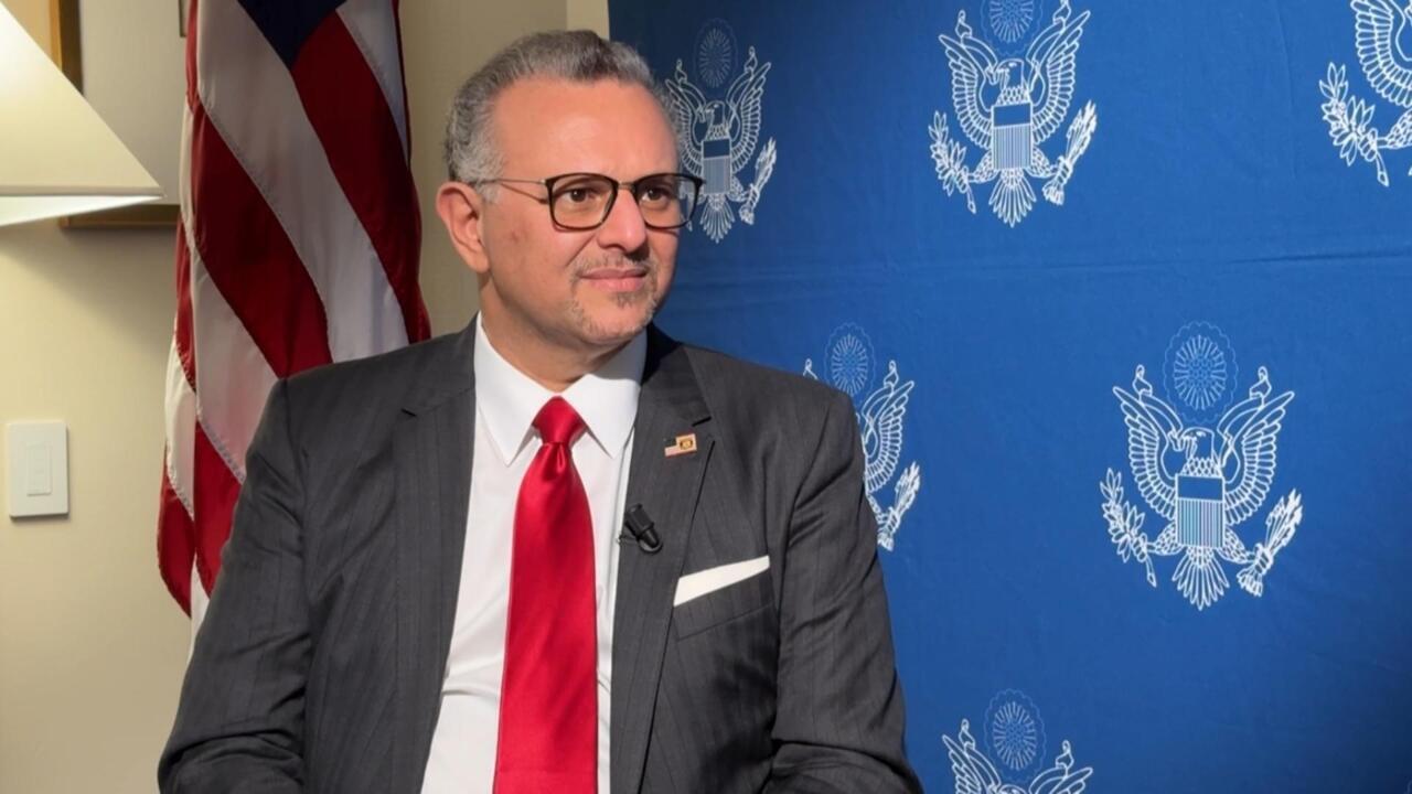 A man in a dark suit and red tie stands before U.S. and blue emblem-patterned flags.