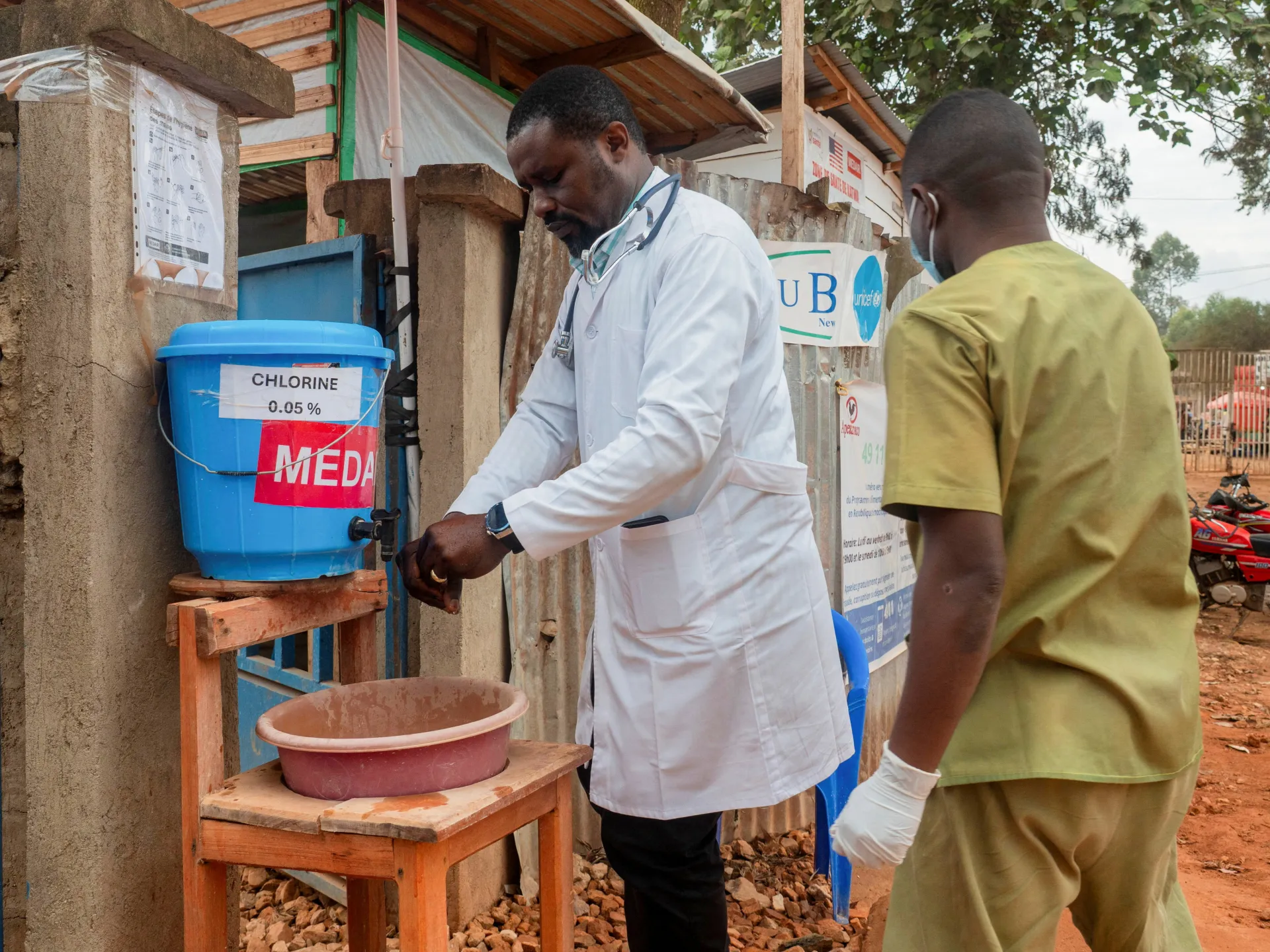 A health worker in protective clothing uses a chlorine handwashing station beside a clinic.