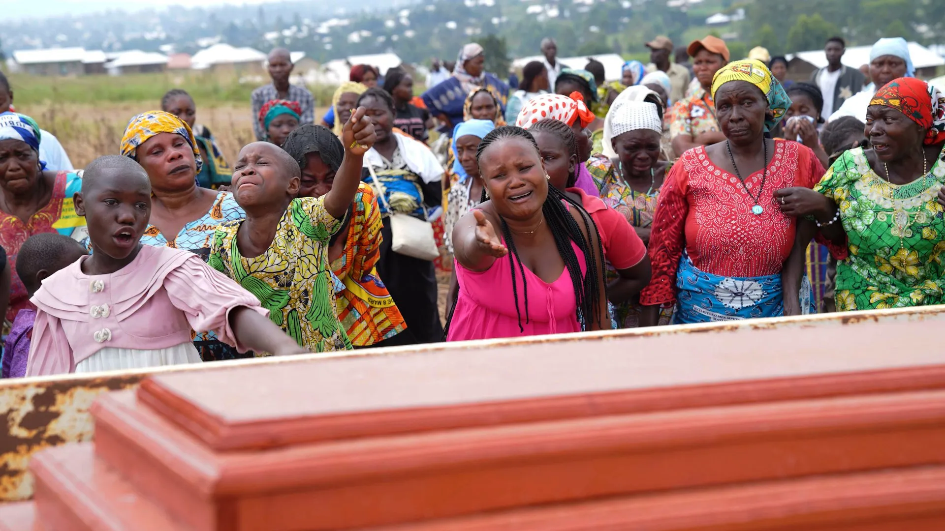 Mourners gather around a reddish coffin as a child and woman cry in the foreground.
