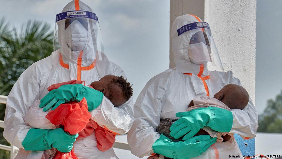 Two healthcare workers in protective suits and face shields hold infants wearing medical gloves.