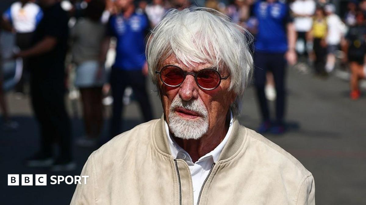 Older white-haired man wearing red-tinted glasses and beige jacket outdoors among blurred people