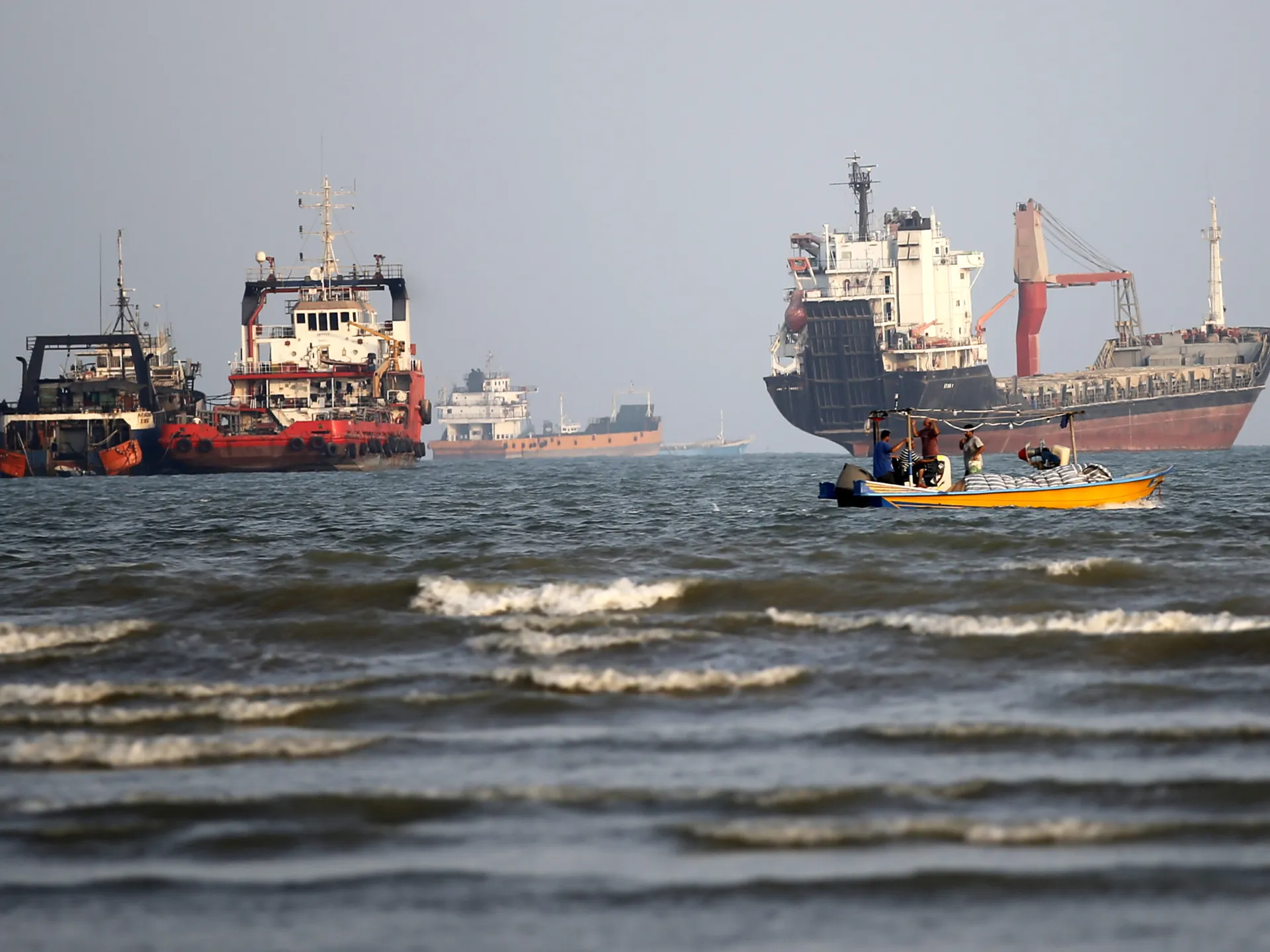 A small yellow boat carrying people passes large cargo ships on choppy water under a hazy sky.
