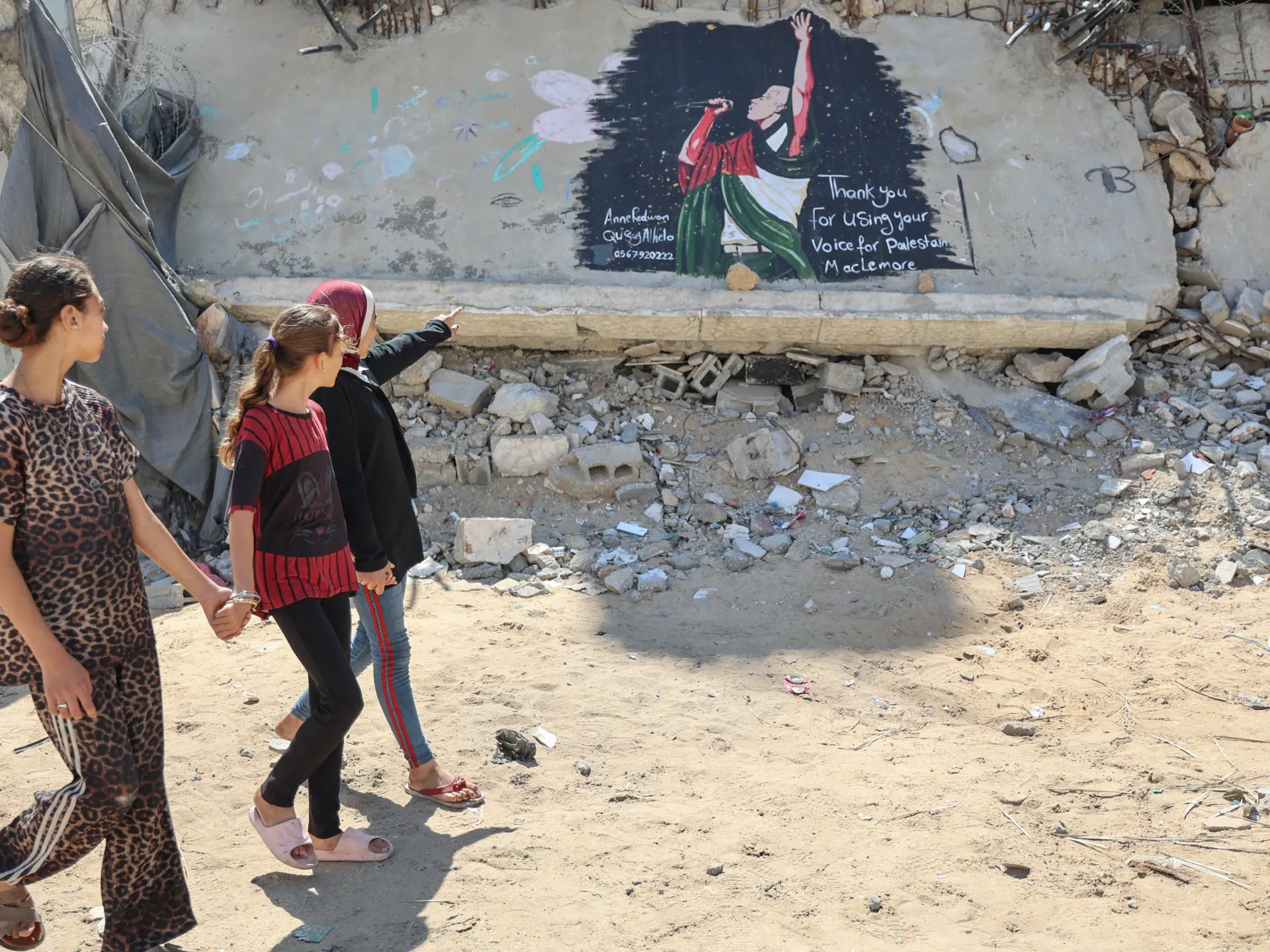 Three women walk past a Palestine-themed mural amid rubble and damaged buildings.
