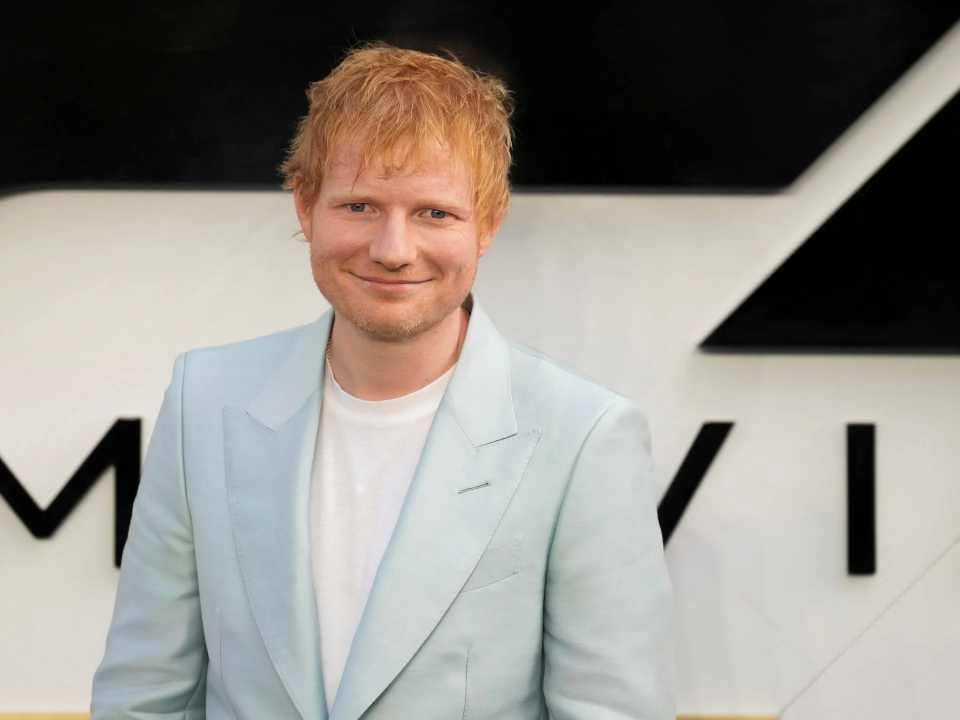 Red-haired man in a pale blue suit jacket stands before a black-and-white backdrop.