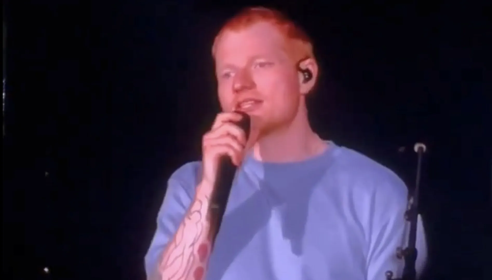 Singer holding a microphone and performing onstage under dim purple lighting.