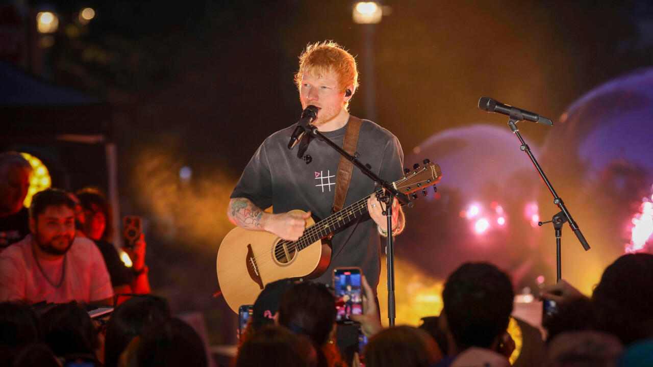 A red-haired singer plays acoustic guitar and sings into a microphone before a concert crowd.