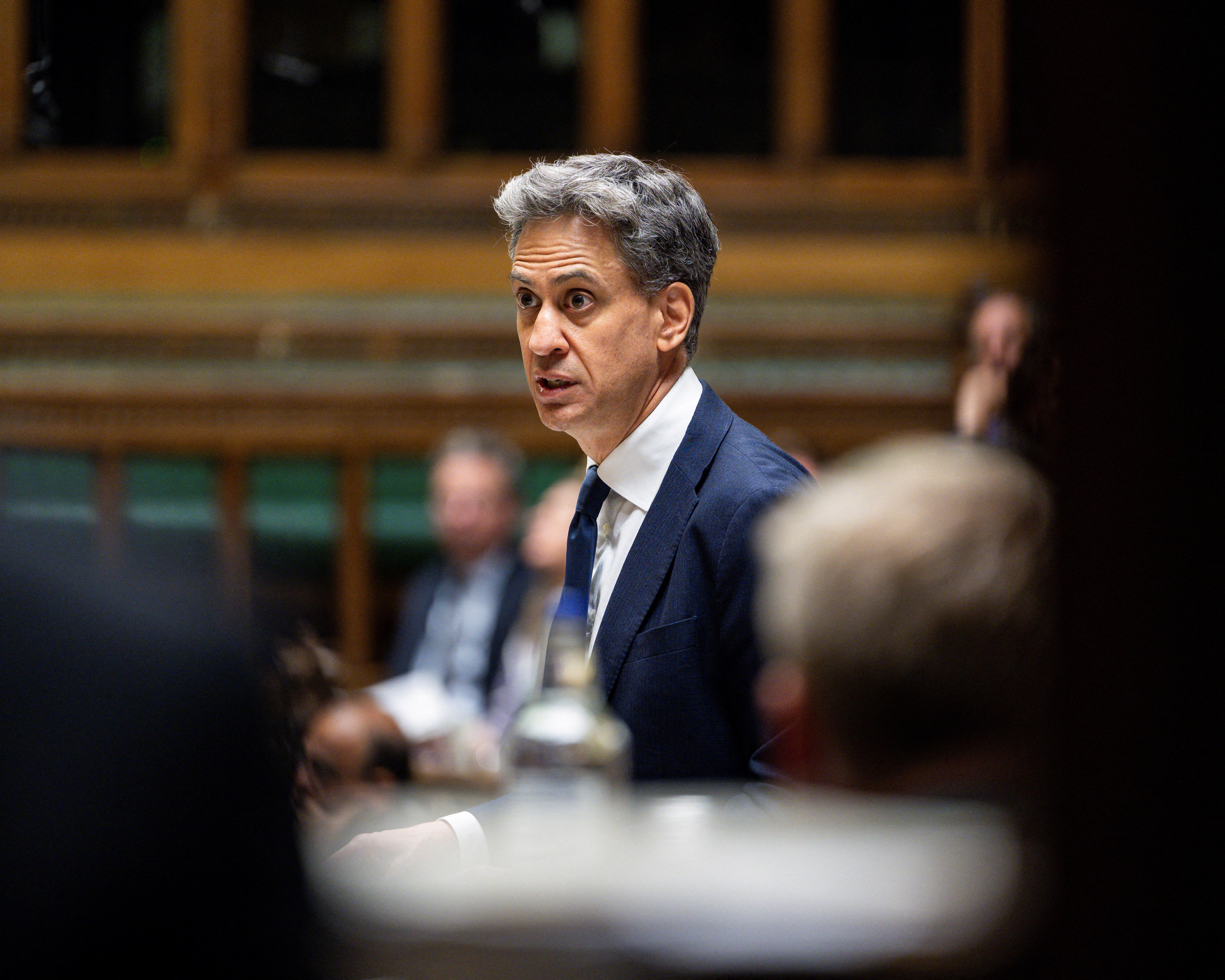 Gray-haired man in a dark suit speaks amid blurred figures in a wood-paneled chamber.