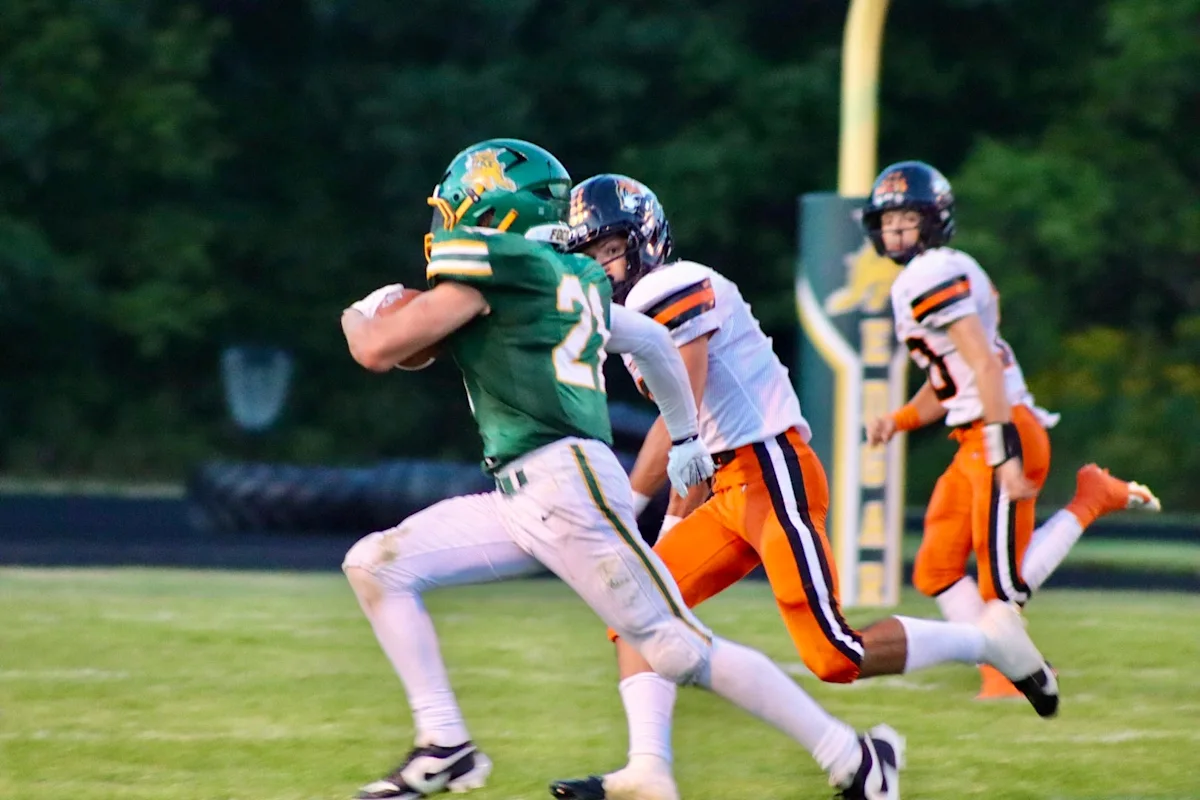 Football player in green runs with the ball as two defenders in orange and white pursue him