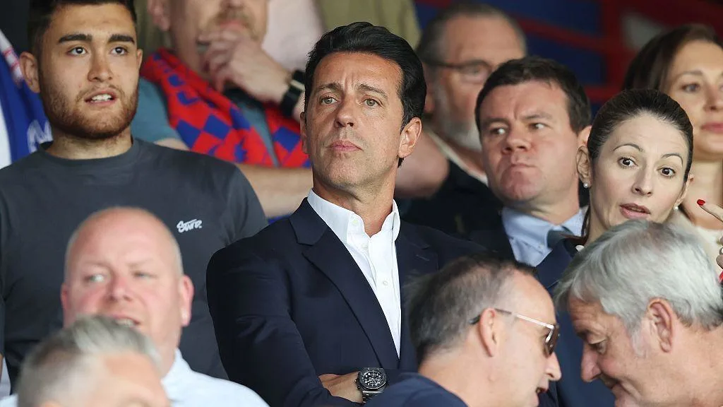 Suited man seated among spectators at a football match, surrounded by other fans