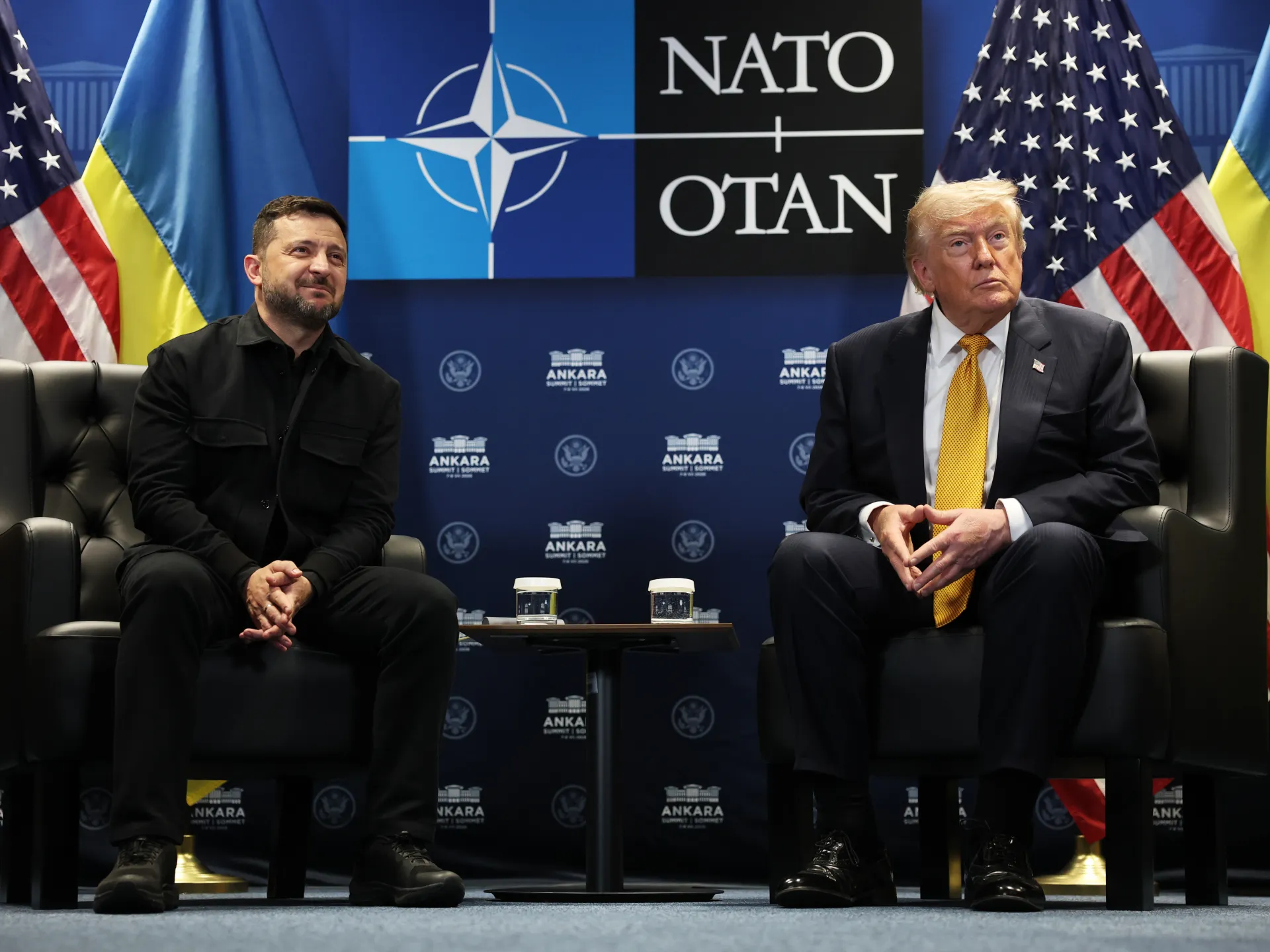 Two political leaders sit in armchairs beneath NATO signage, with Ukrainian and American flags behind them.