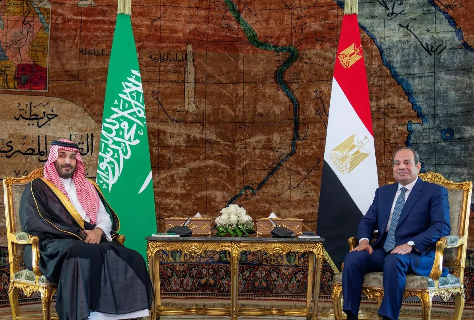 Two officials seated beside Saudi and Egyptian flags, with a historic map displayed behind them.