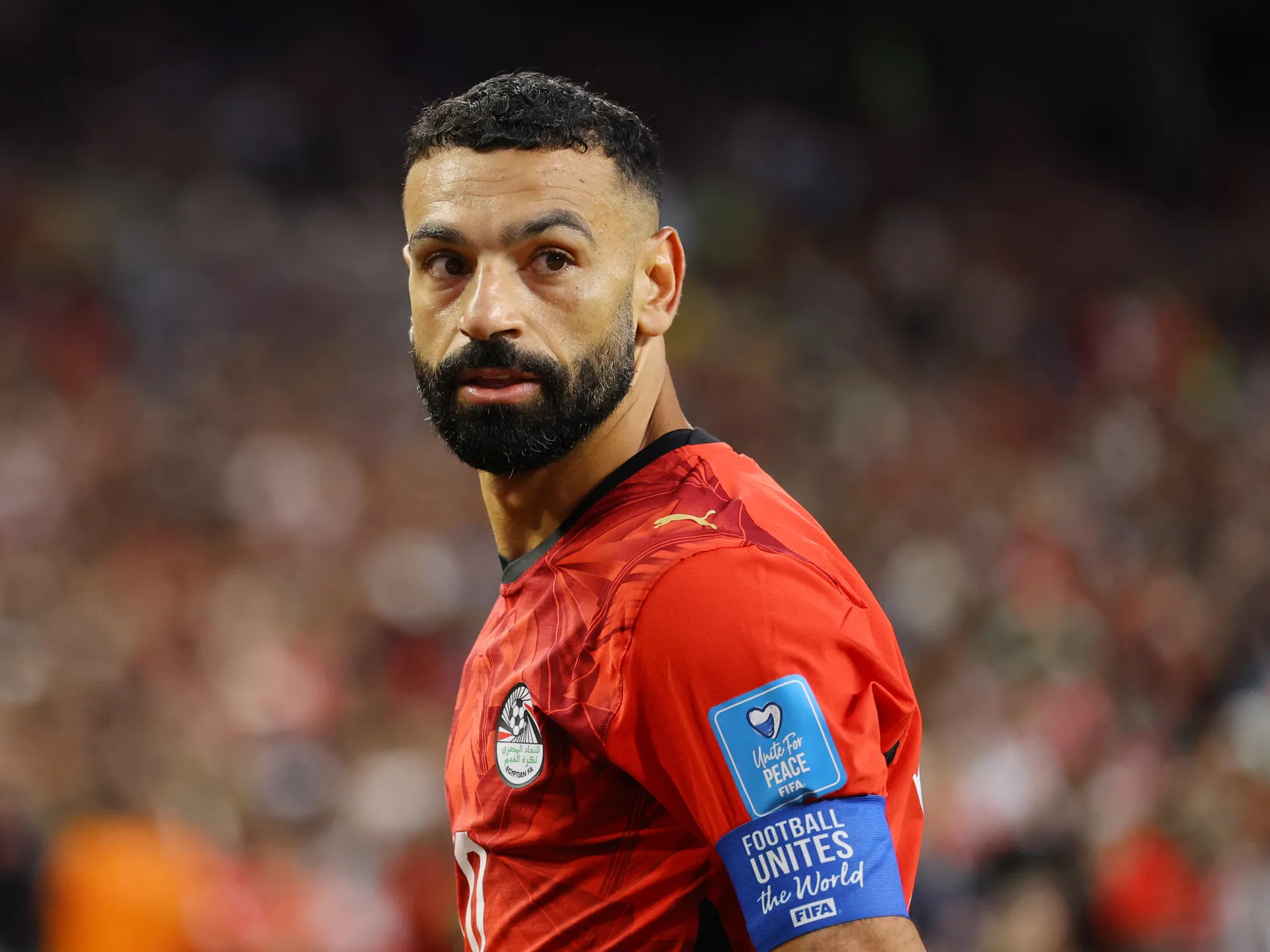 Footballer in a red Egypt jersey and blue captain’s armband looks toward the camera on the pitch.