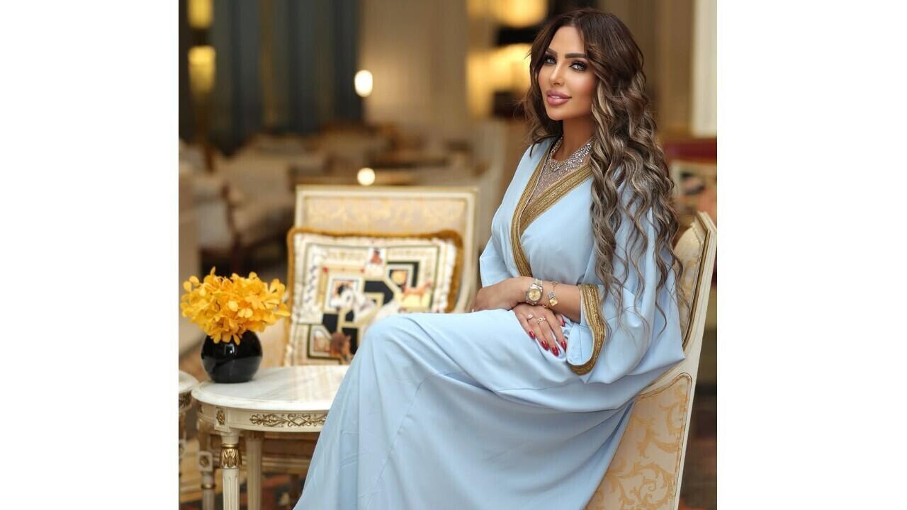 Woman in a light blue, gold-trimmed dress seated indoors beside flowers and ornate furniture.