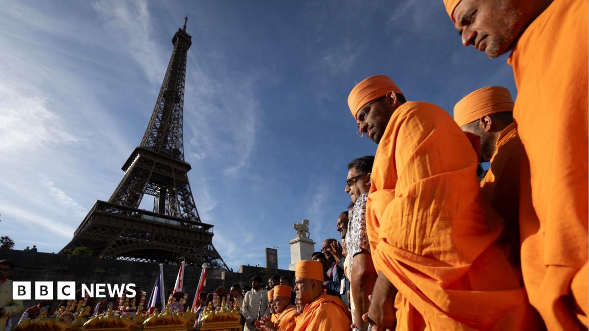 Men in orange robes stand beside a flower-covered display near the Eiffel Tower.