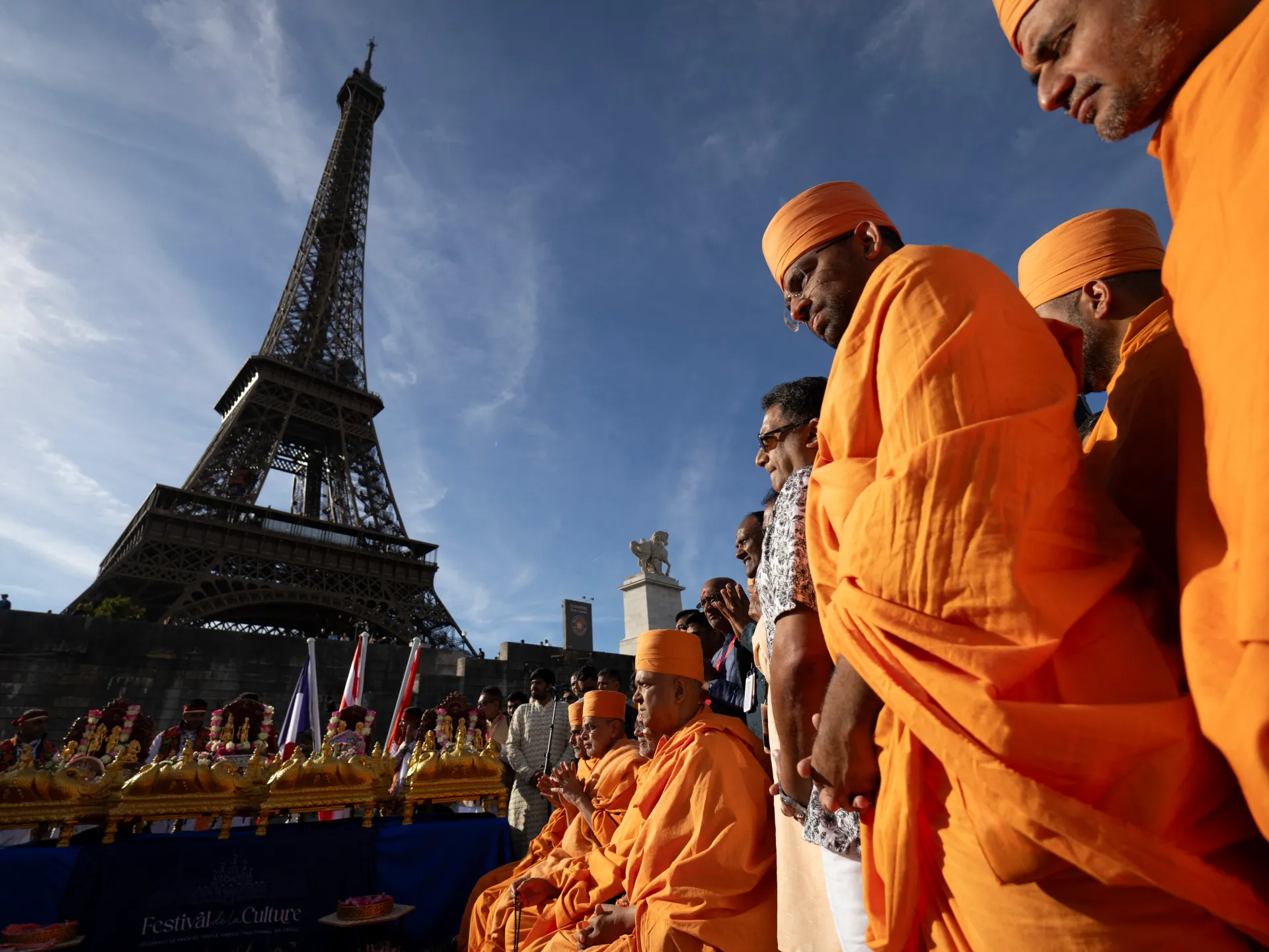 Orange-robed people gather beside the Eiffel Tower under a clear blue sky.