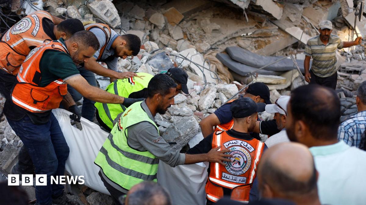 Rescuers in orange and green vests carry a white body bag through rubble from a collapsed building.