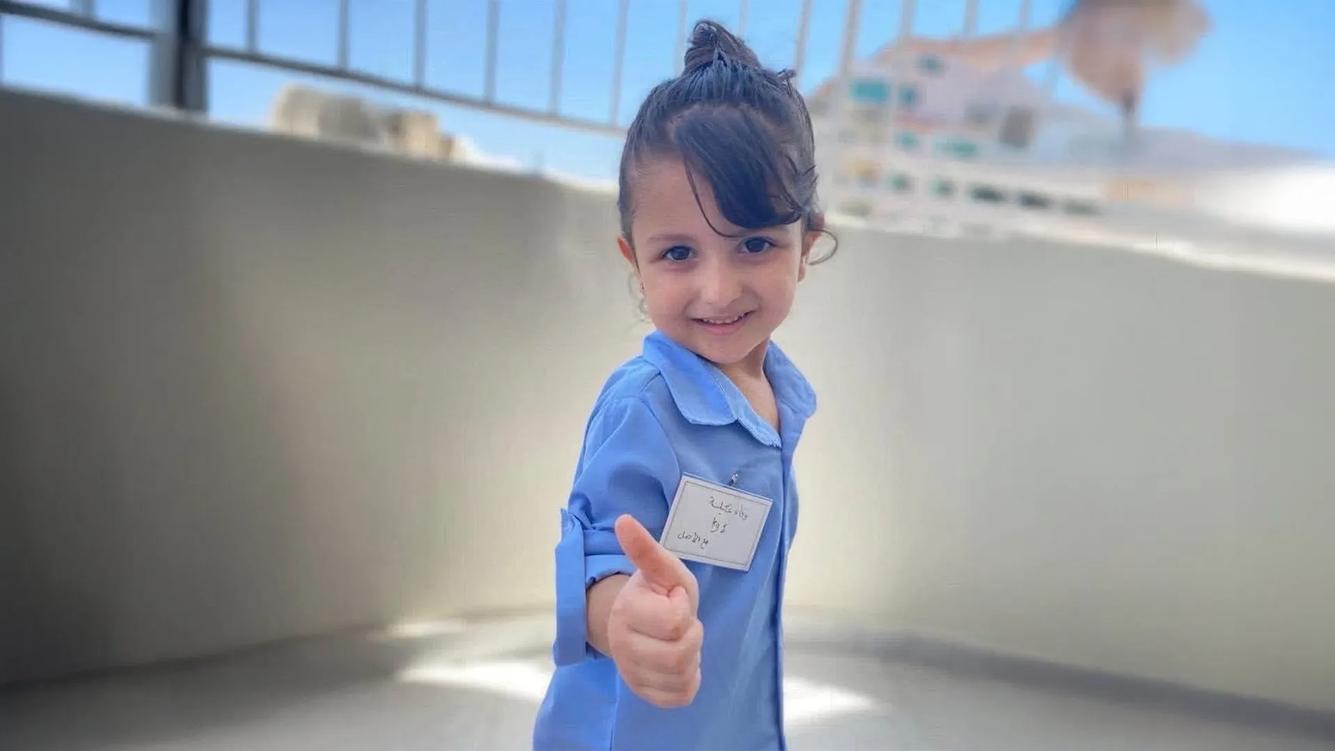 Young girl in blue school uniform smiling and giving a thumbs-up indoors