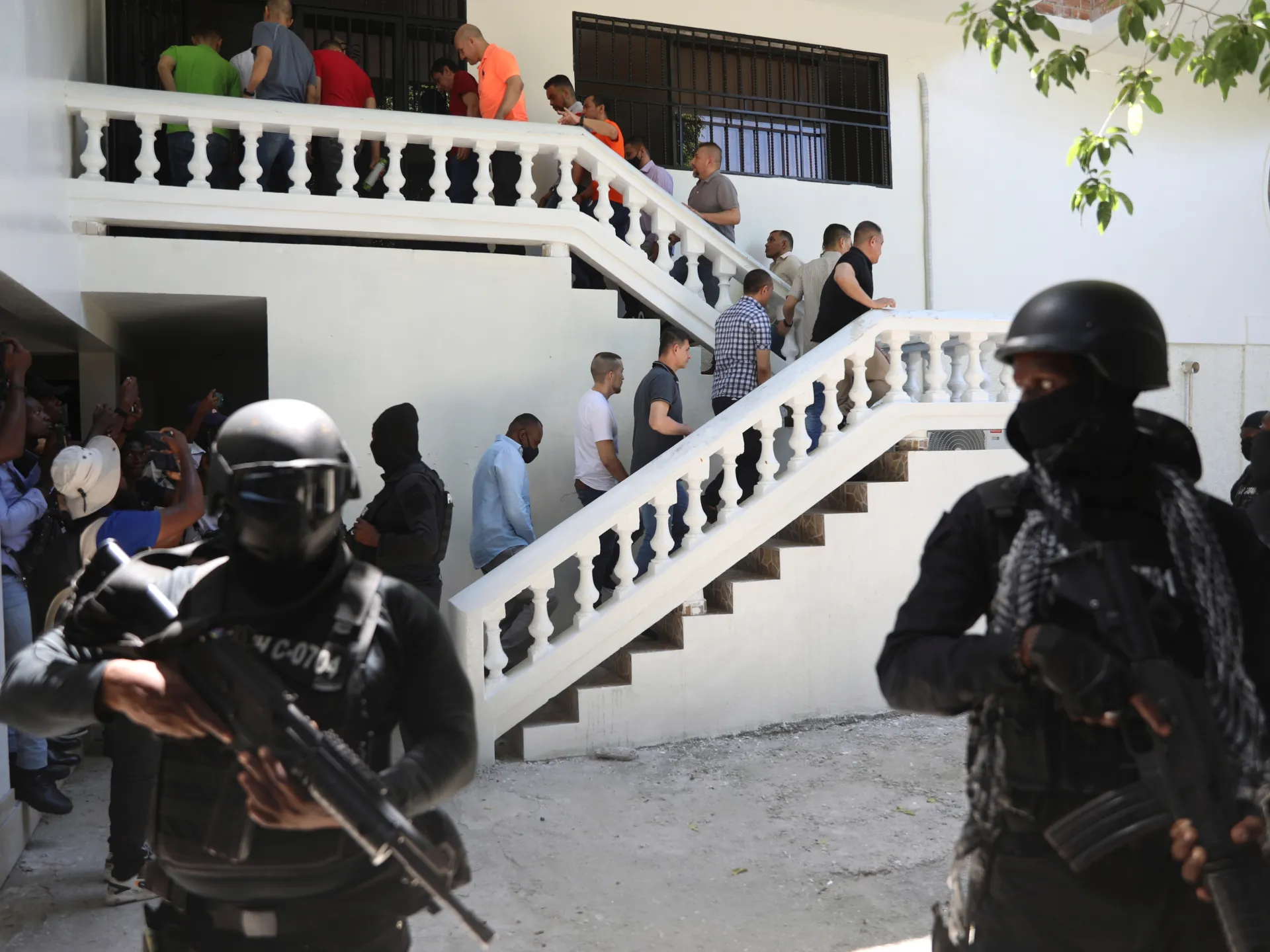 Heavily armed officers stand near a white staircase as civilians gather inside a building.