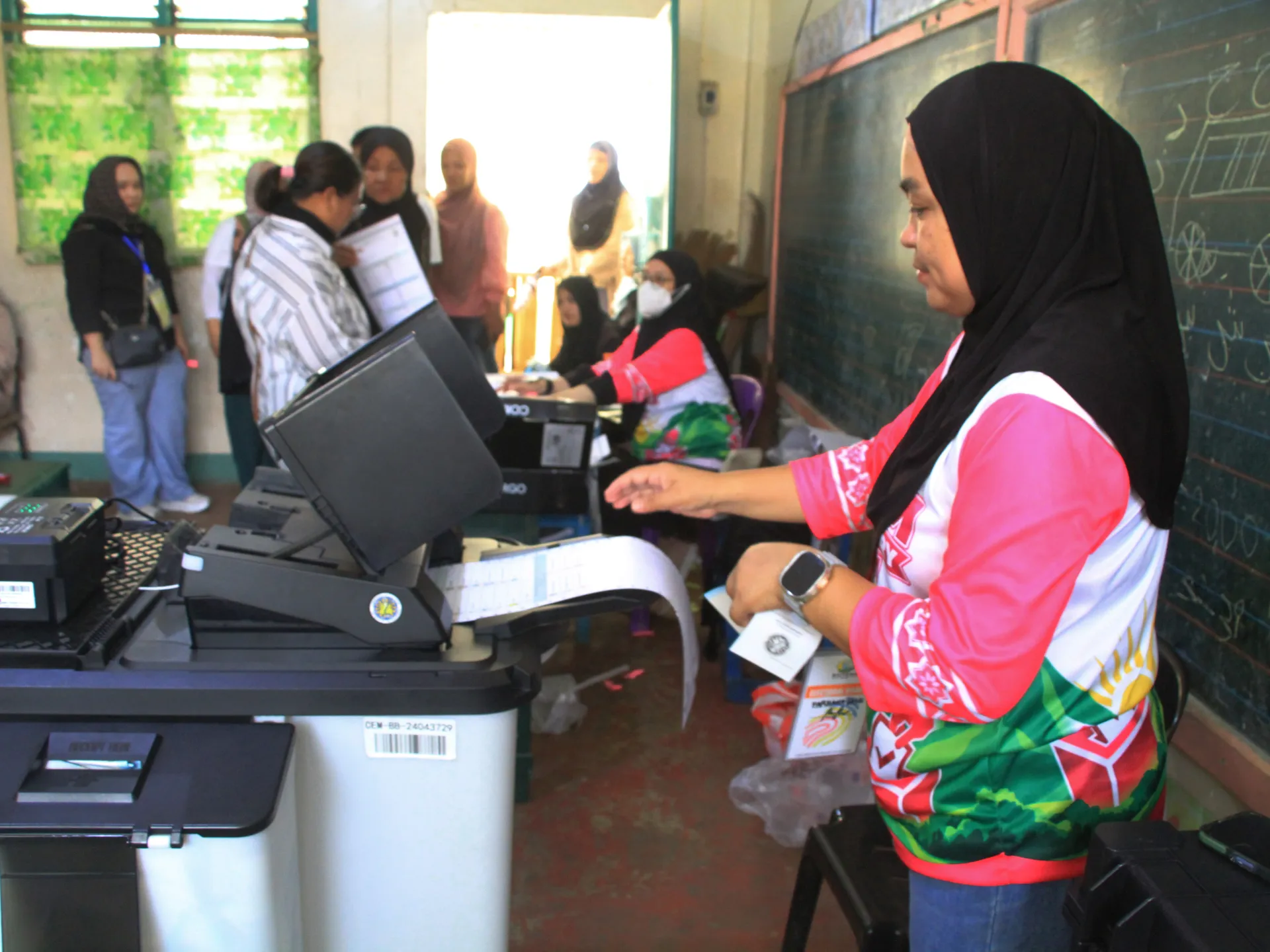 Woman in colorful shirt operates election equipment inside a crowded polling room.
