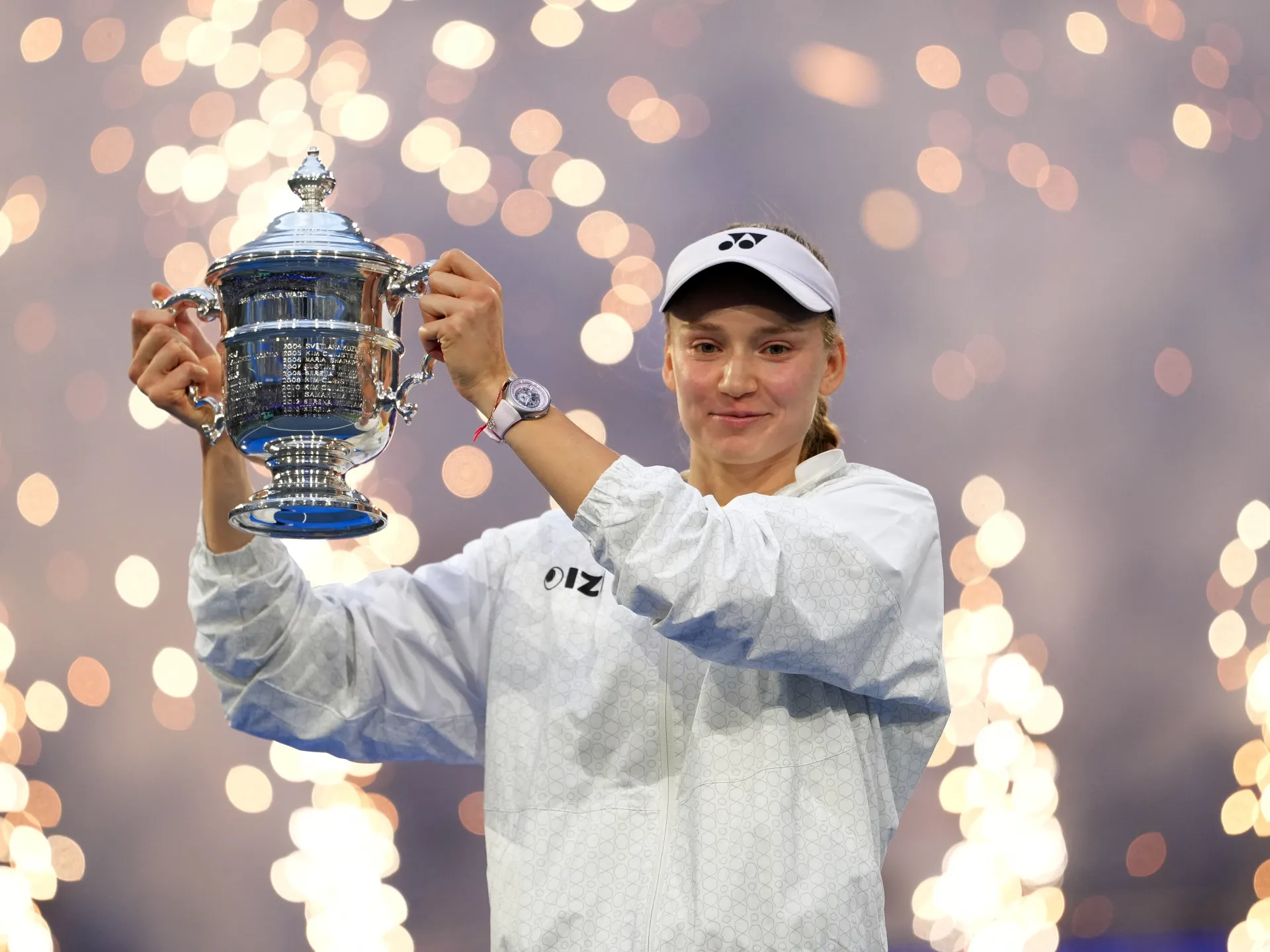 Tennis player in white visor and jacket raises a silver championship trophy amid celebratory lights.