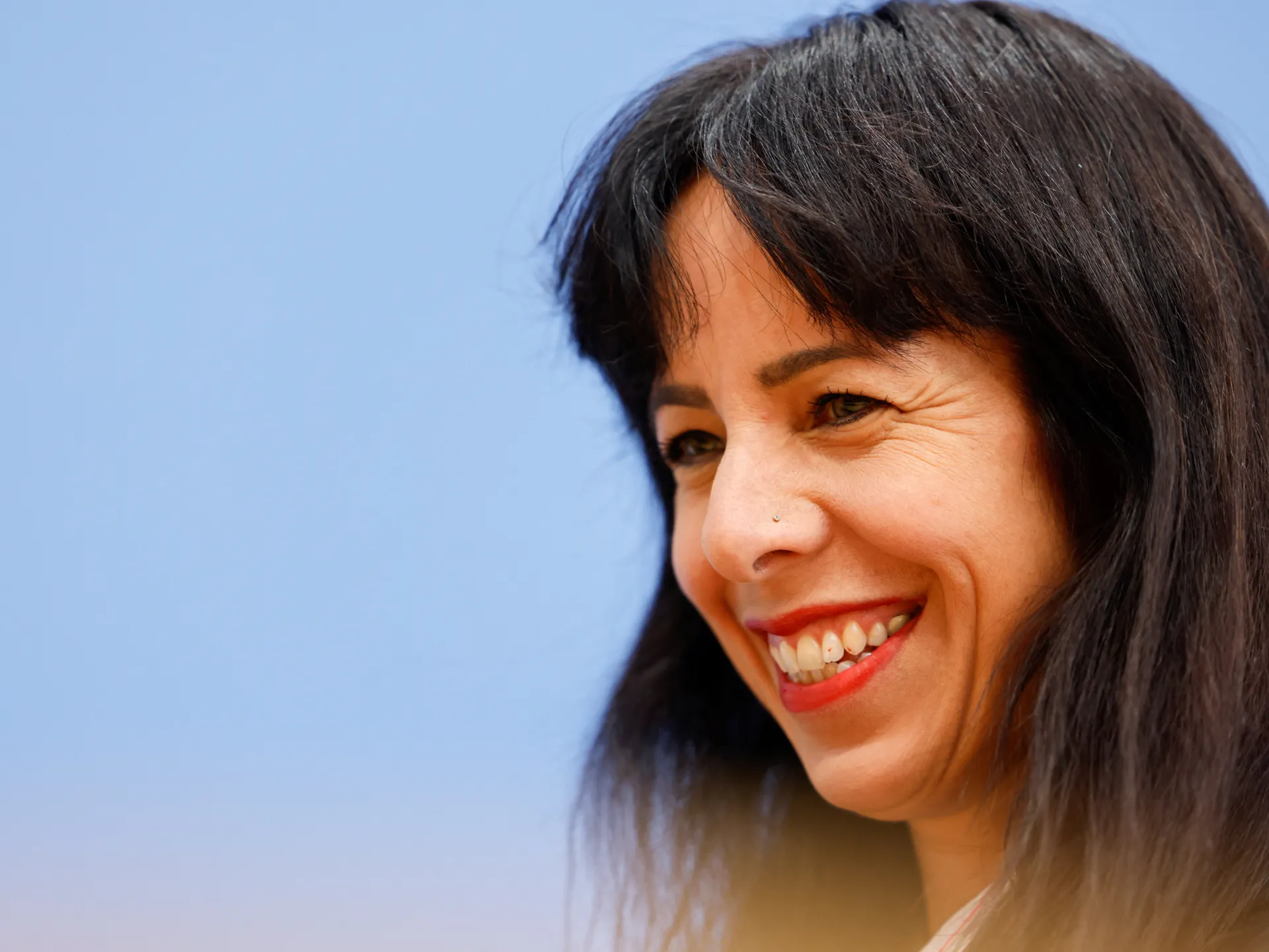 Smiling woman with dark hair and red lipstick photographed in close-up against a pale blue background