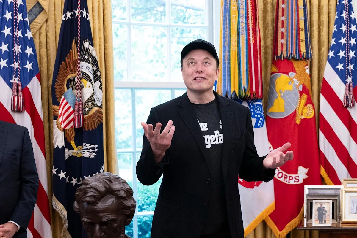Elon Musk gestures while standing among American and military flags in an ornate room.