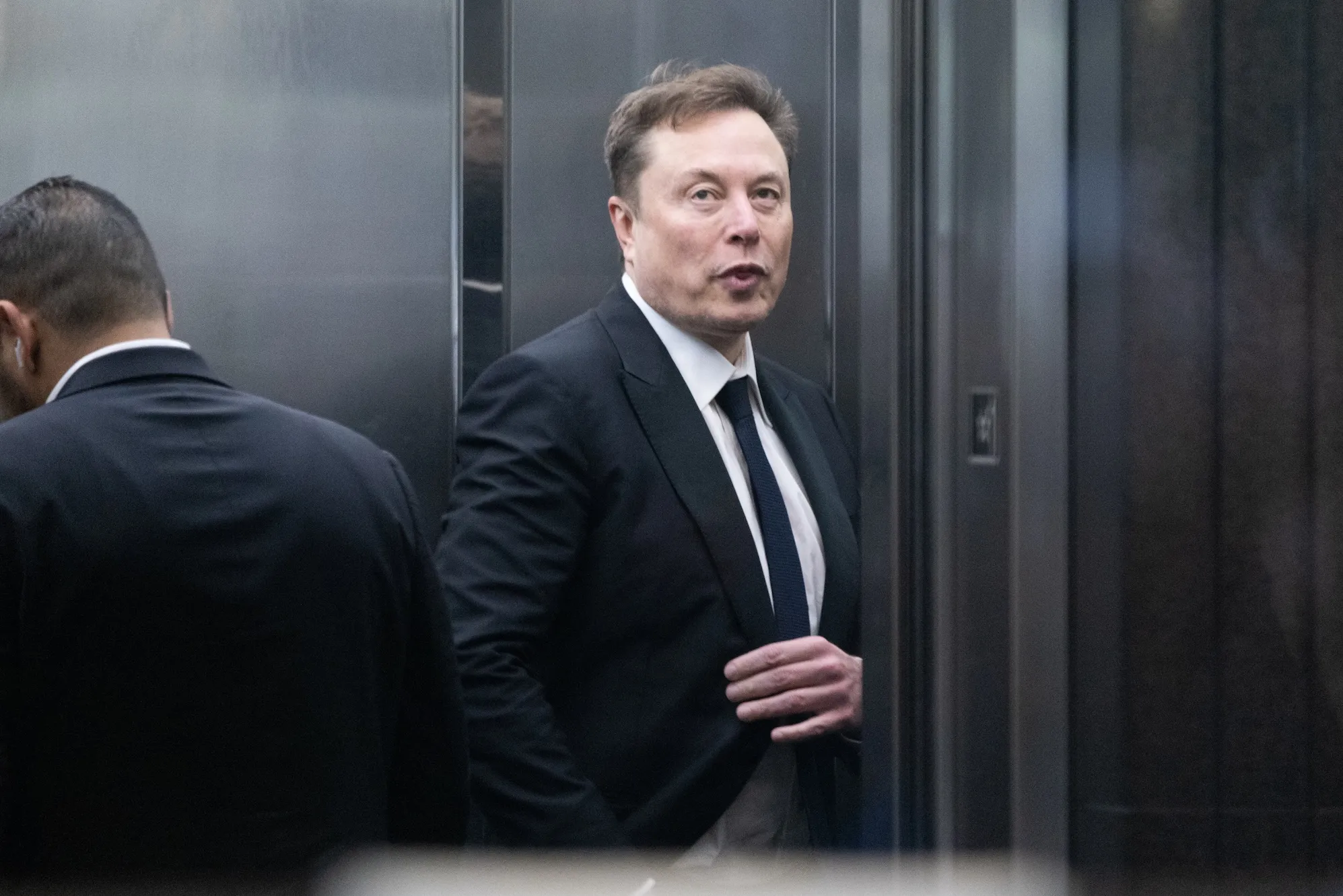 Man in dark suit stands beside an elevator, adjusting his tie while another man faces away.