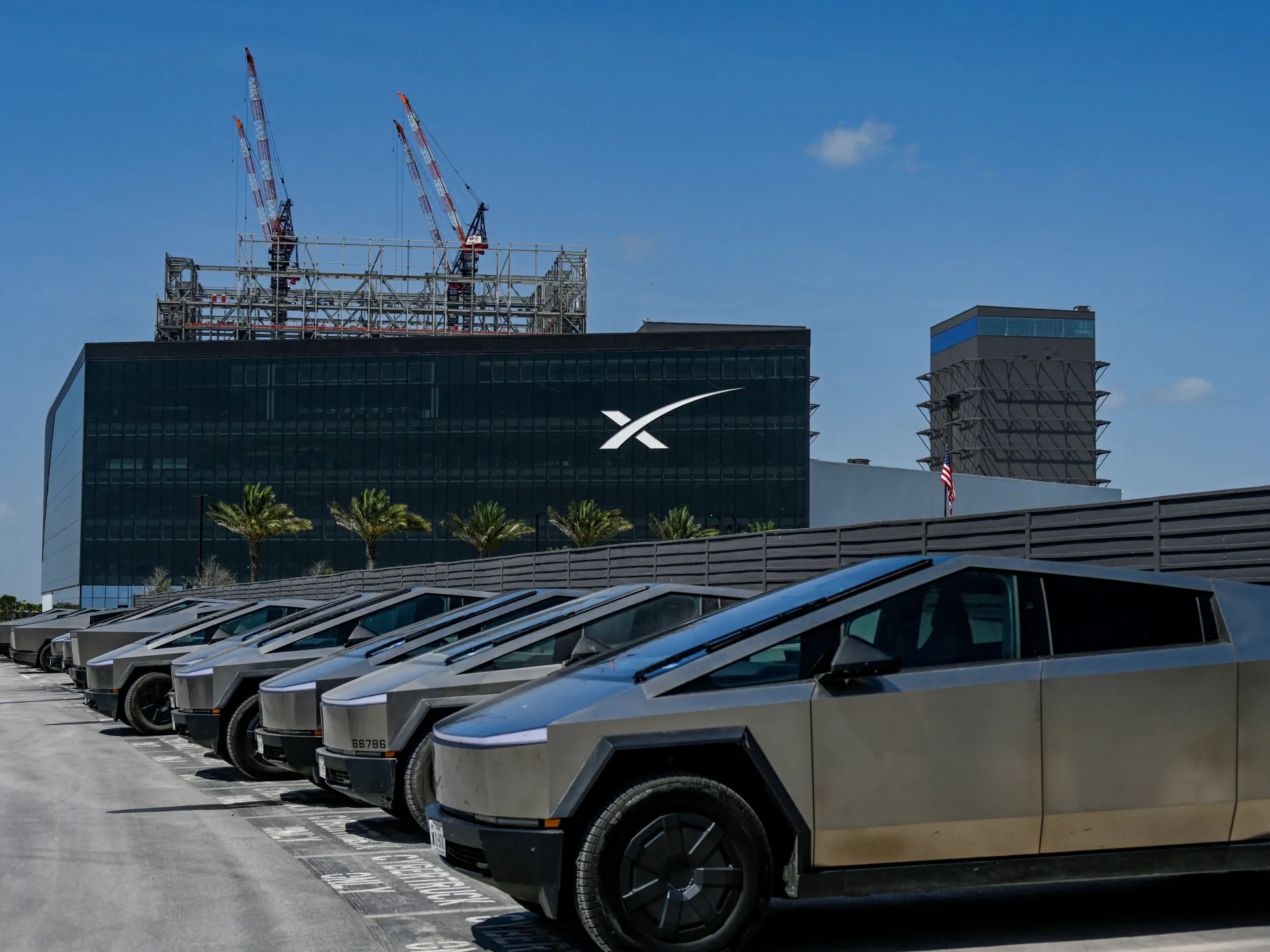 Rows of Cybertrucks parked outside an X-branded building with construction cranes overhead