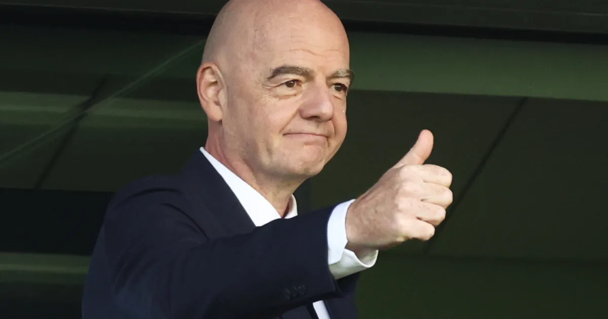 Bald man in dark suit gives a thumbs-up gesture while seated indoors.