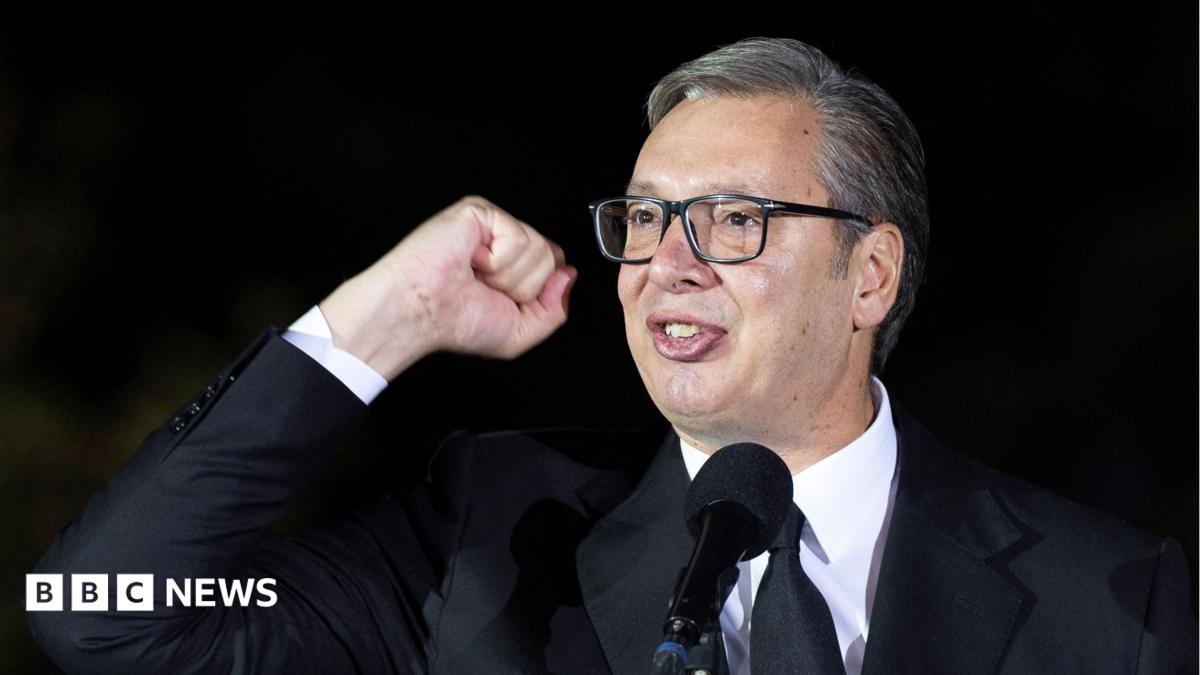 A bespectacled man in a dark suit raises a fist while speaking into a microphone against a dark background.