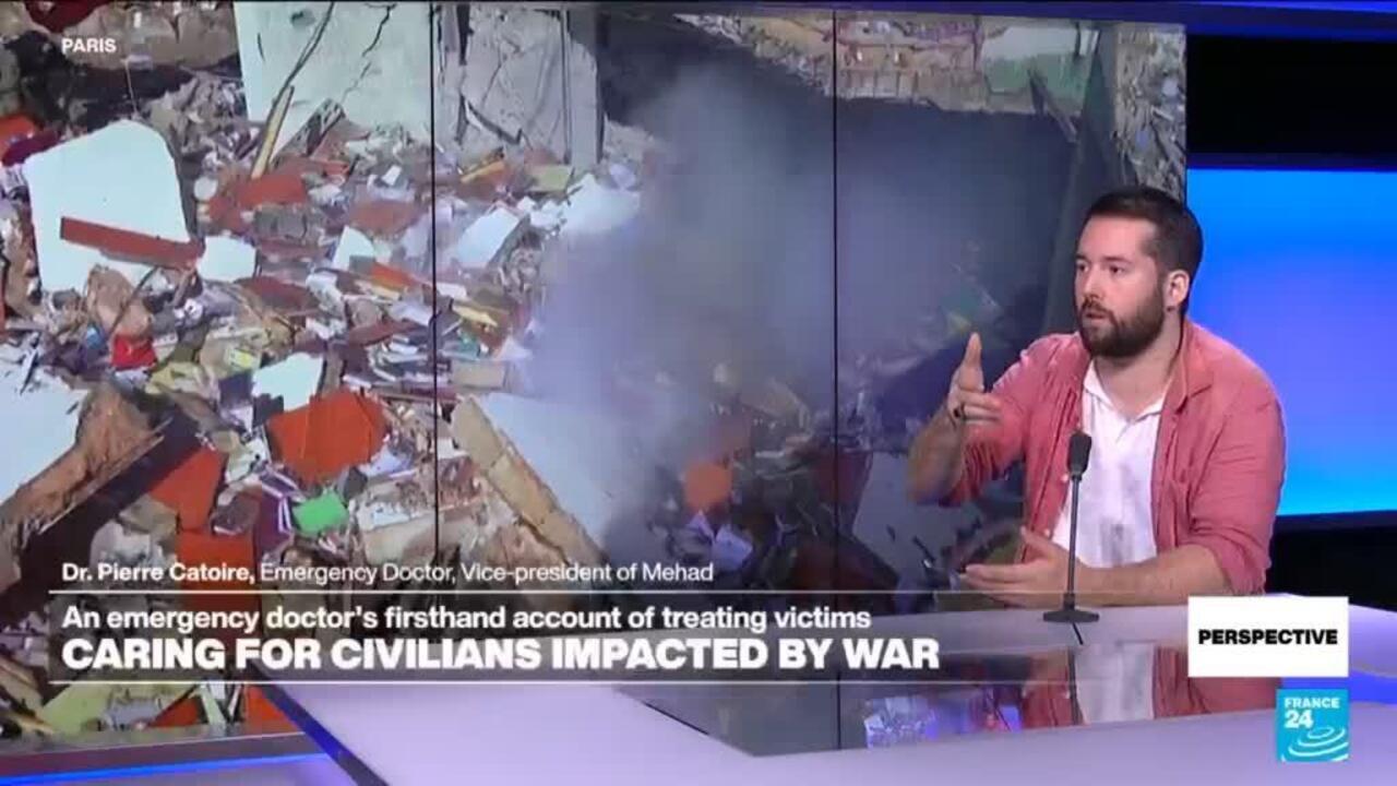 Bearded doctor speaks in a television studio beside footage of heavily damaged buildings and rubble.