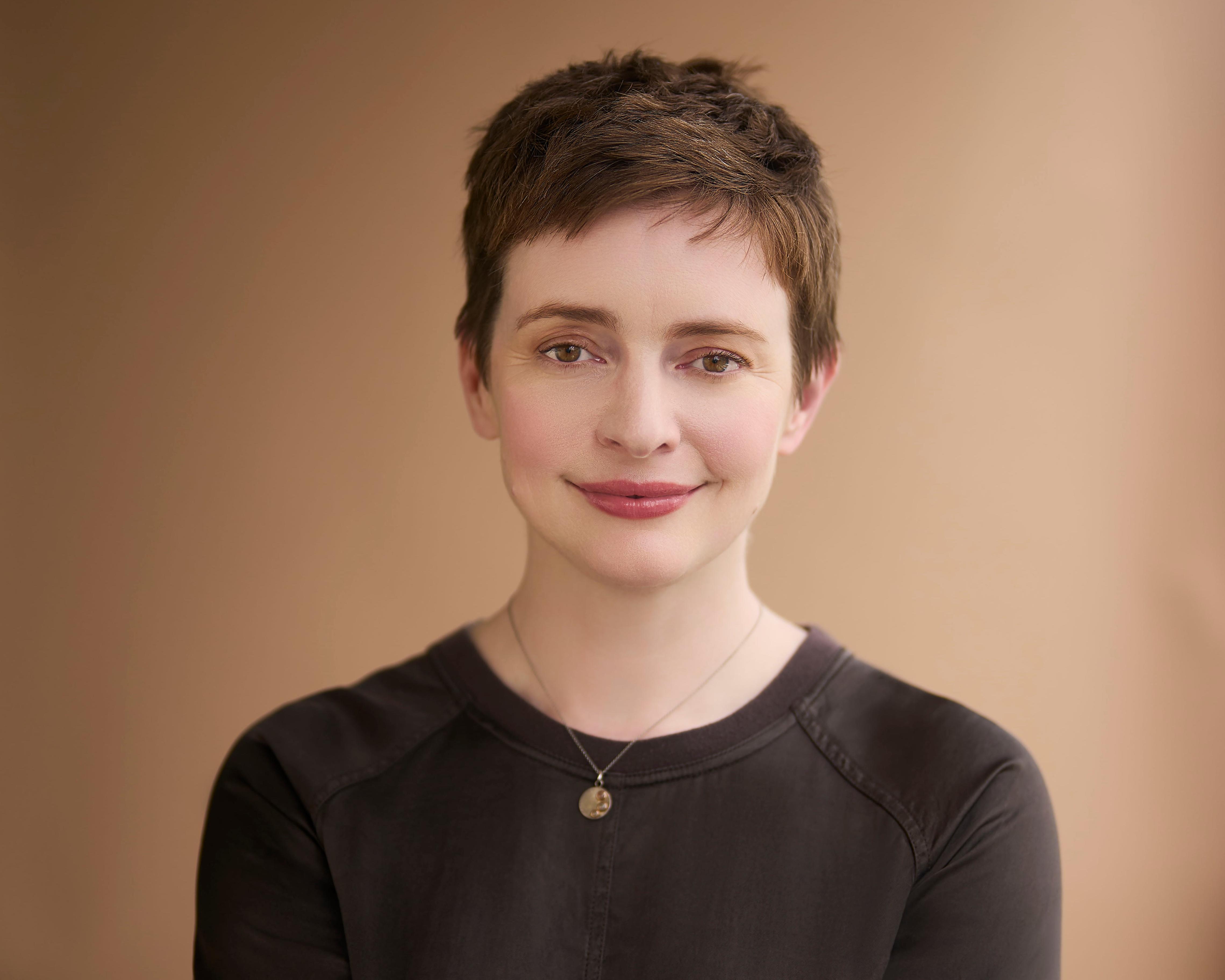 Portrait of a short-haired person wearing a dark top and pendant necklace against a warm beige background.