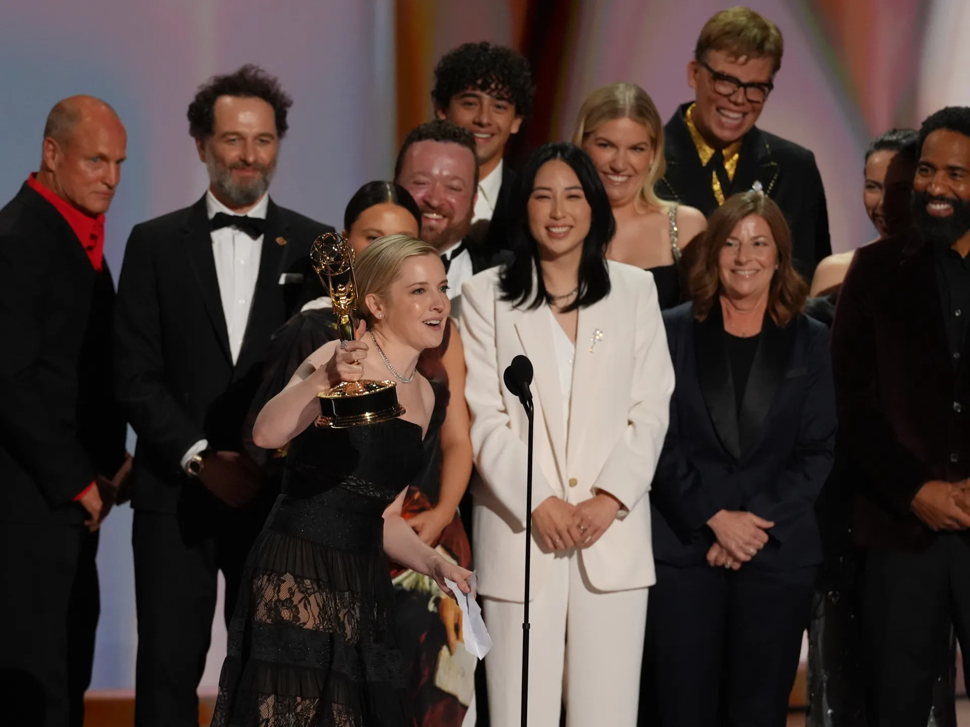 Group of television professionals celebrates onstage as a woman holds an Emmy statuette near a microphone.