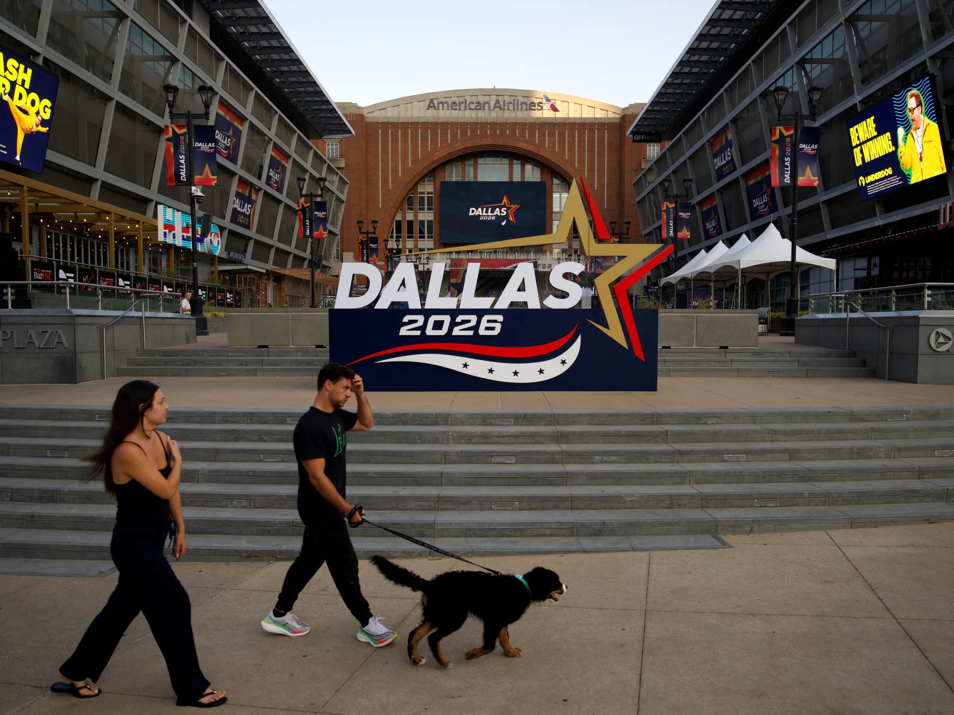 People walk a dog past a Dallas 2026 convention display outside an arena.