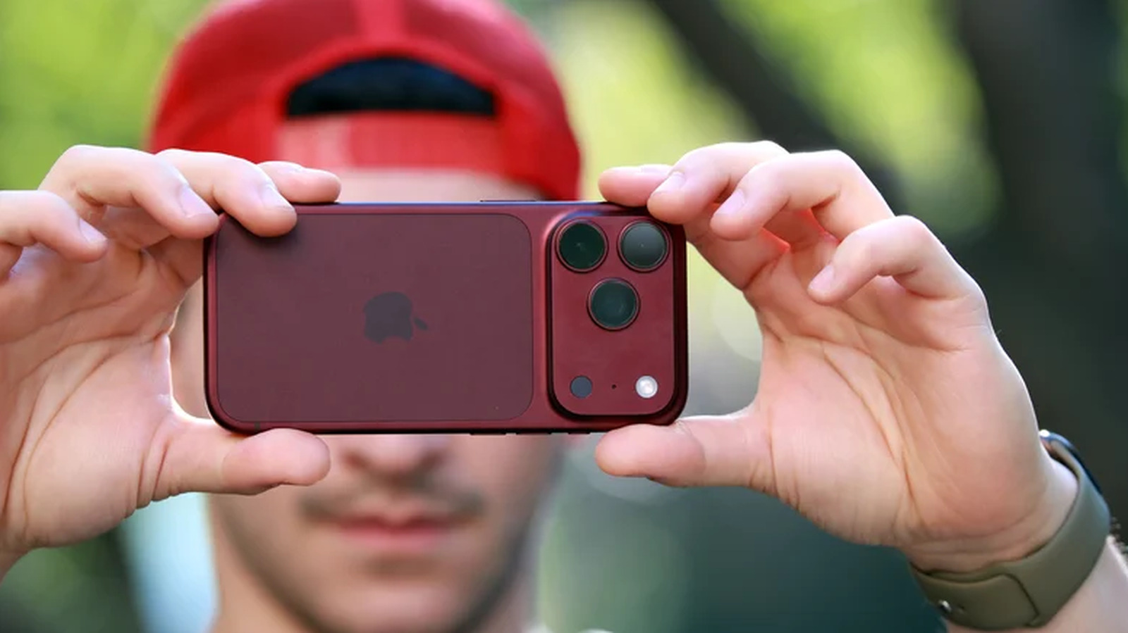 A person in a red cap holds a burgundy smartphone horizontally with its rear cameras facing forward.