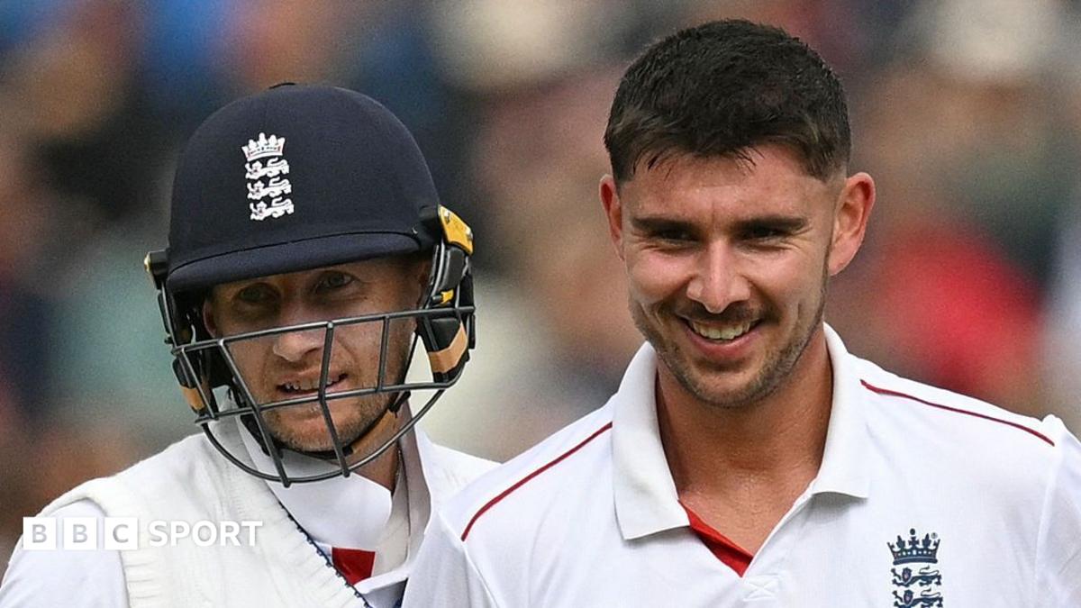 Two England cricketers in white Test uniforms, one helmeted, stand together smiling on the field.