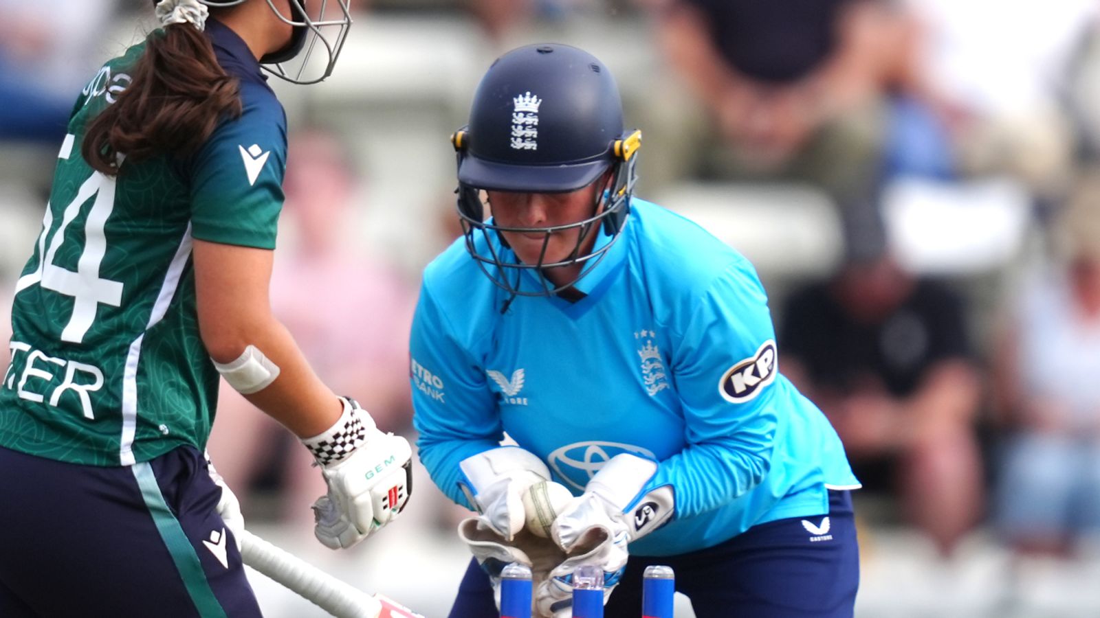 England wicketkeeper crouches behind the stumps as a batter in green approaches during a cricket match.