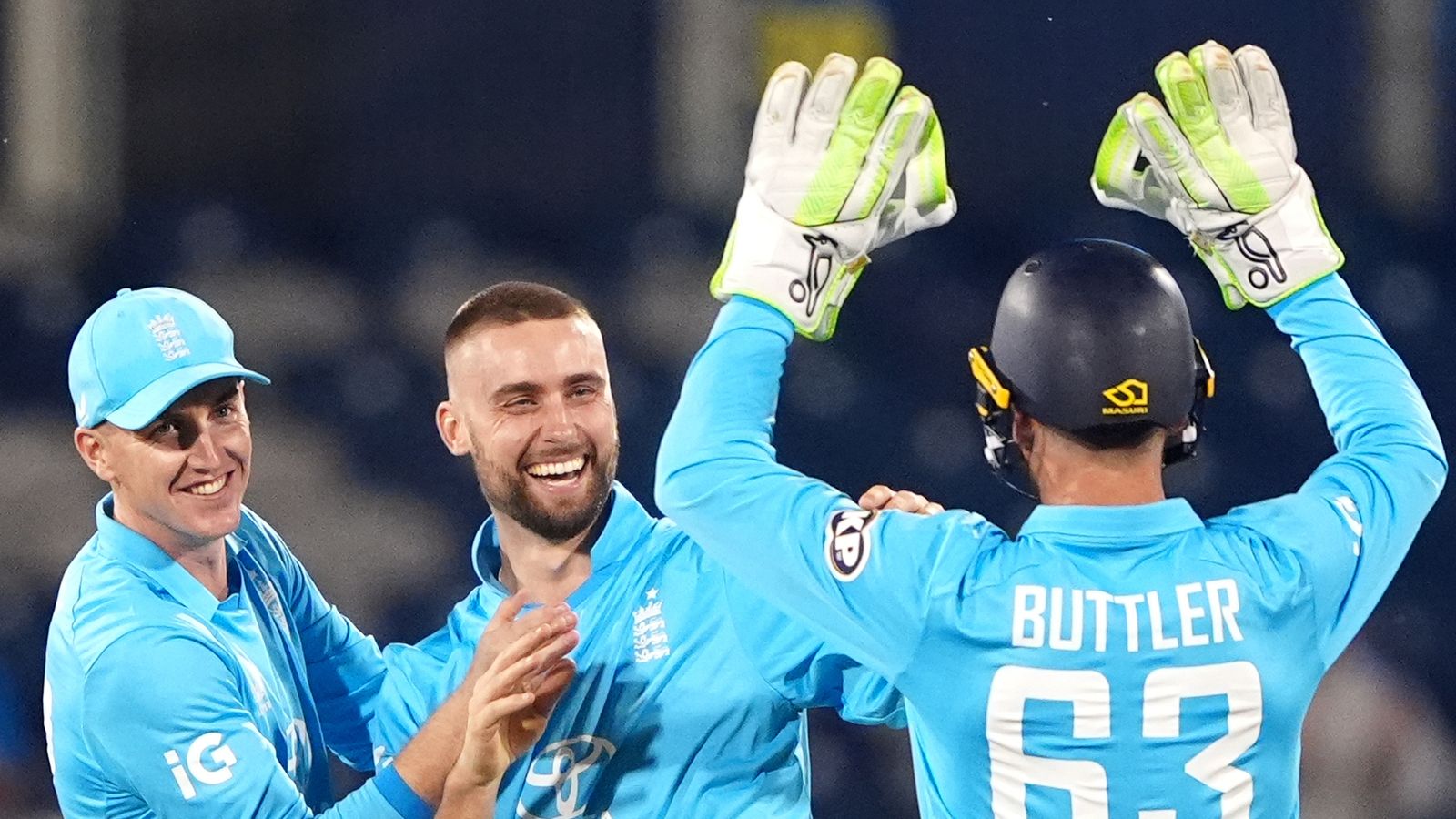 Three England cricketers celebrate as the wicketkeeper raises his gloved hands behind a teammate.