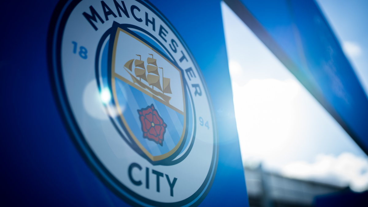 Close-up of the Manchester City crest on a blue surface, with bright stadium lights and sky in the background.