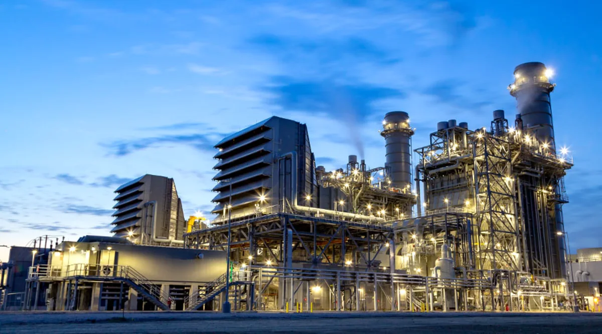 Illuminated industrial power plant with tall stacks and steel structures beneath a blue evening sky.