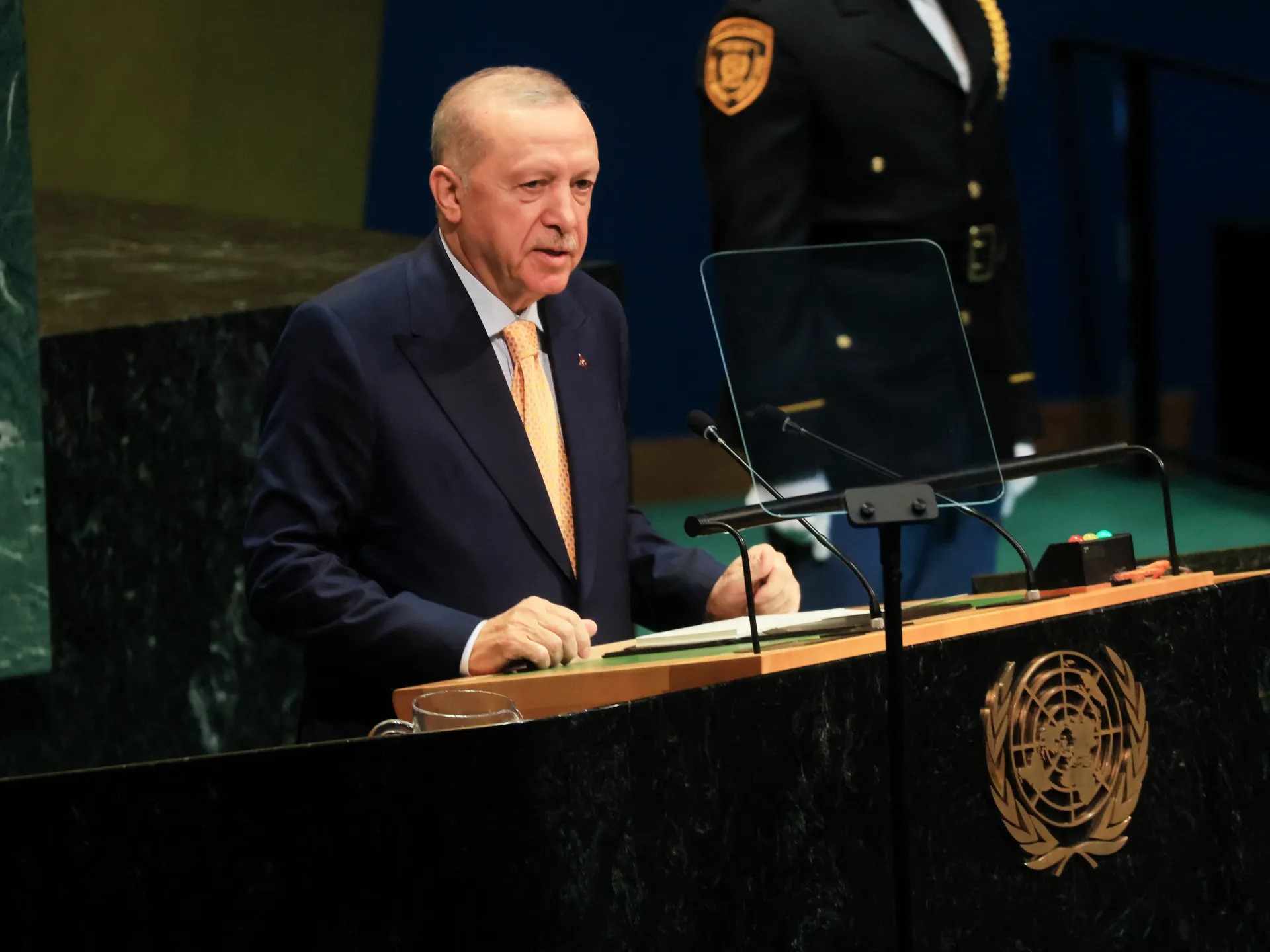 Man in dark suit and orange tie speaking at a lectern with transparent teleprompters