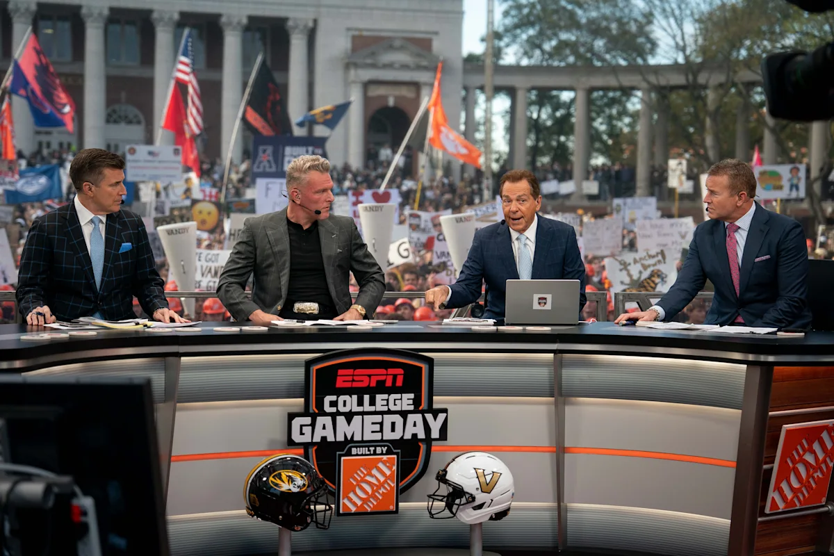 Four television analysts sit at an ESPN College GameDay desk before a cheering crowd.