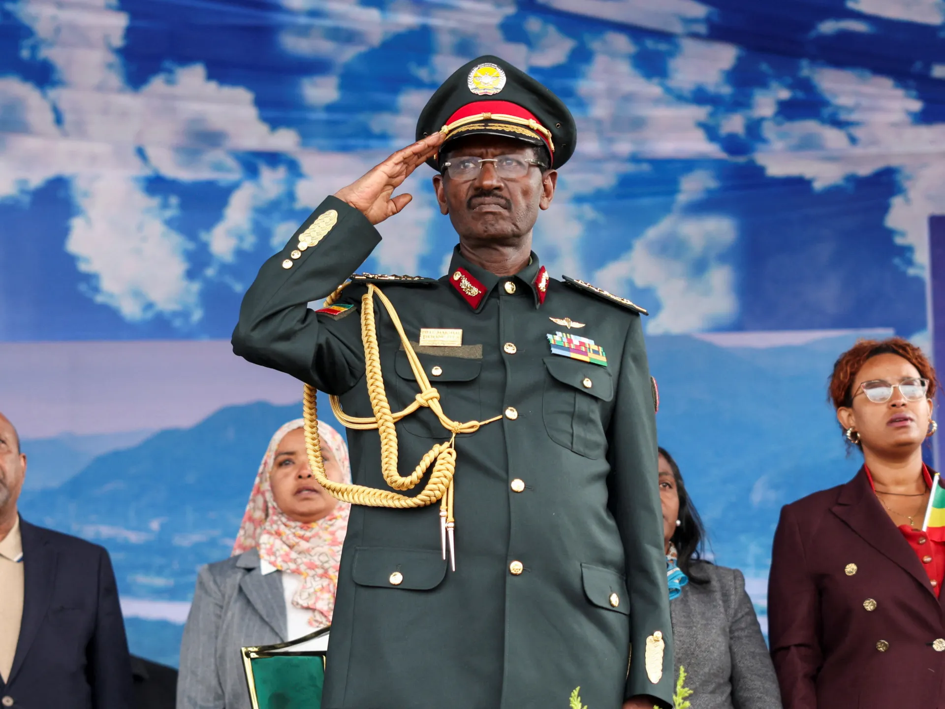 Uniformed officer salutes in front of a blue backdrop, with officials standing behind him.
