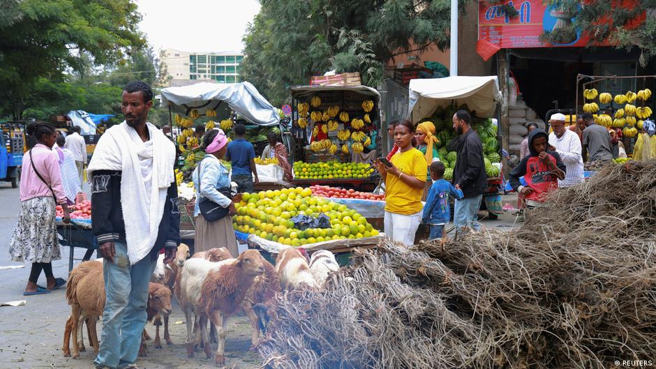 People shop at a busy outdoor market with fruit stalls, livestock, and bundles of branches.