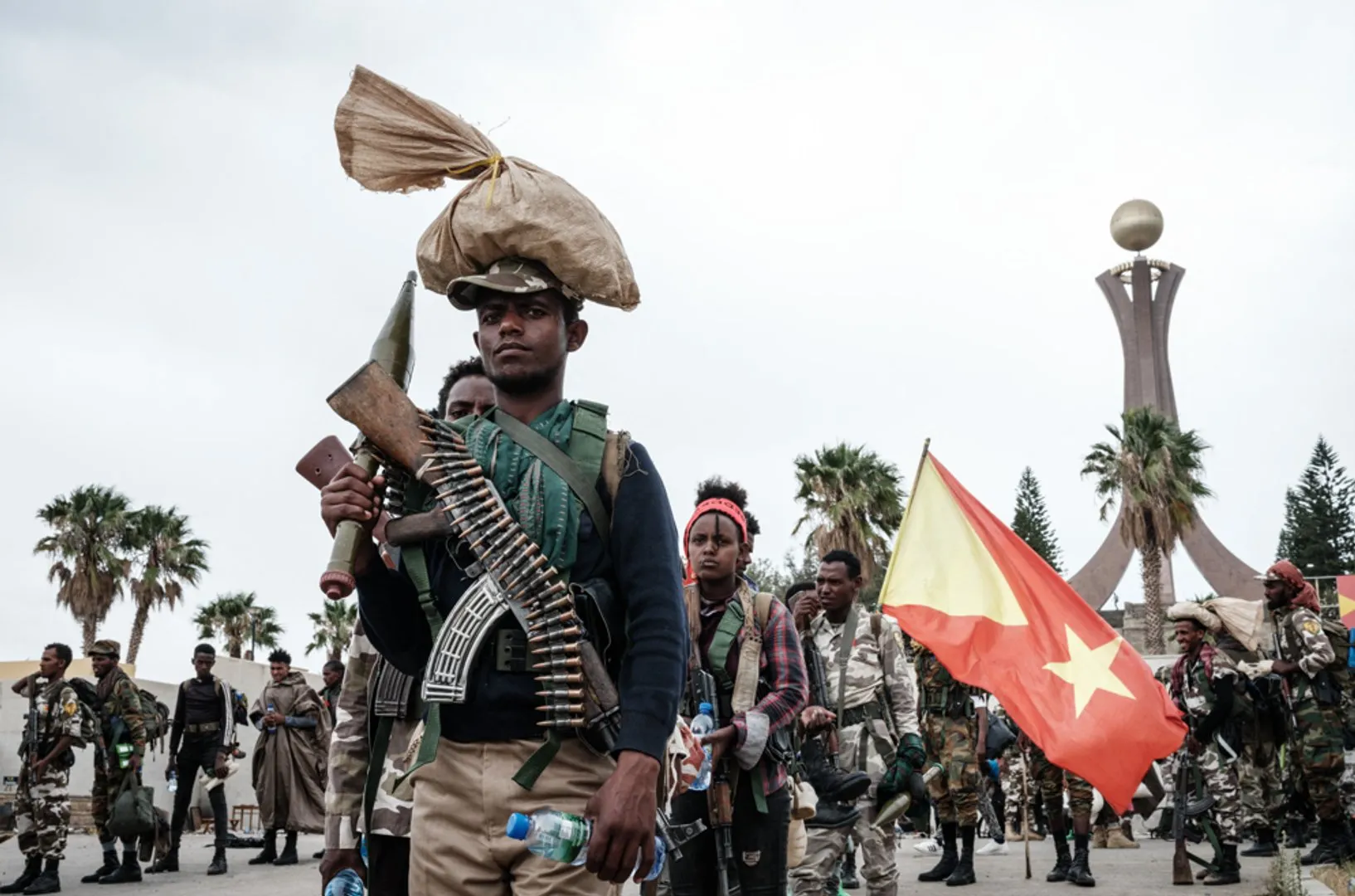 Armed men gather in a public square, one carrying a rifle and ammunition beside a red star flag.