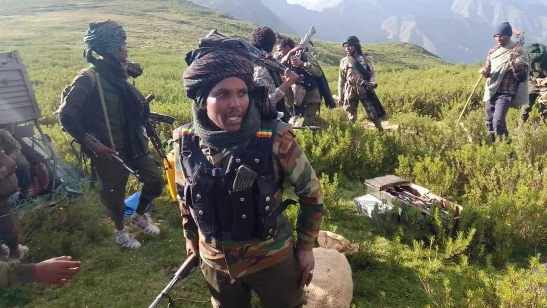 Armed men in mixed clothing stand in a grassy mountain area beside equipment and a vehicle.