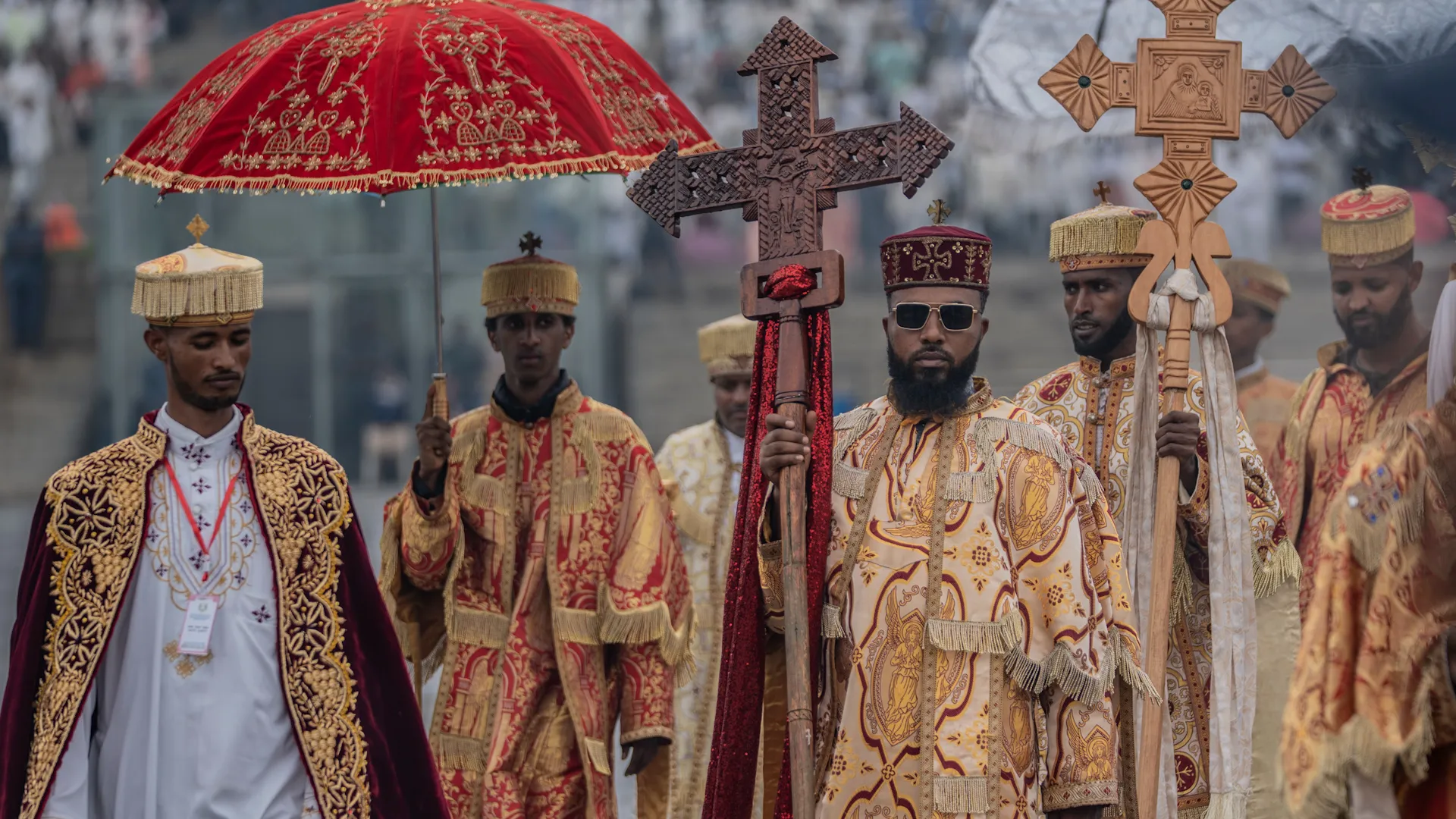 Ethiopian Orthodox clergy in embroidered robes carry crosses and a red ceremonial umbrella in a procession.