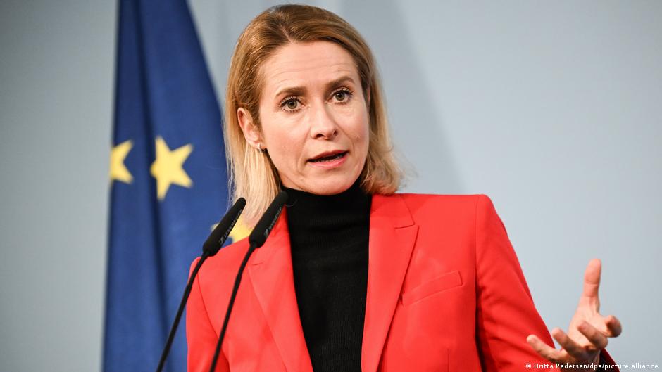 Blonde woman in red blazer speaking at a podium with European Union flags behind her
