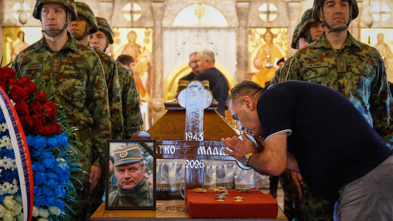 Man bows beside a coffin as uniformed soldiers stand nearby in a church filled with flowers.