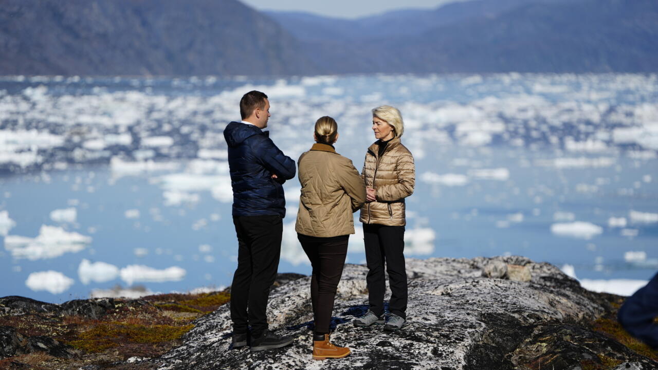 Three people stand on rocky terrain overlooking an ice-dotted fjord and mountainous landscape.